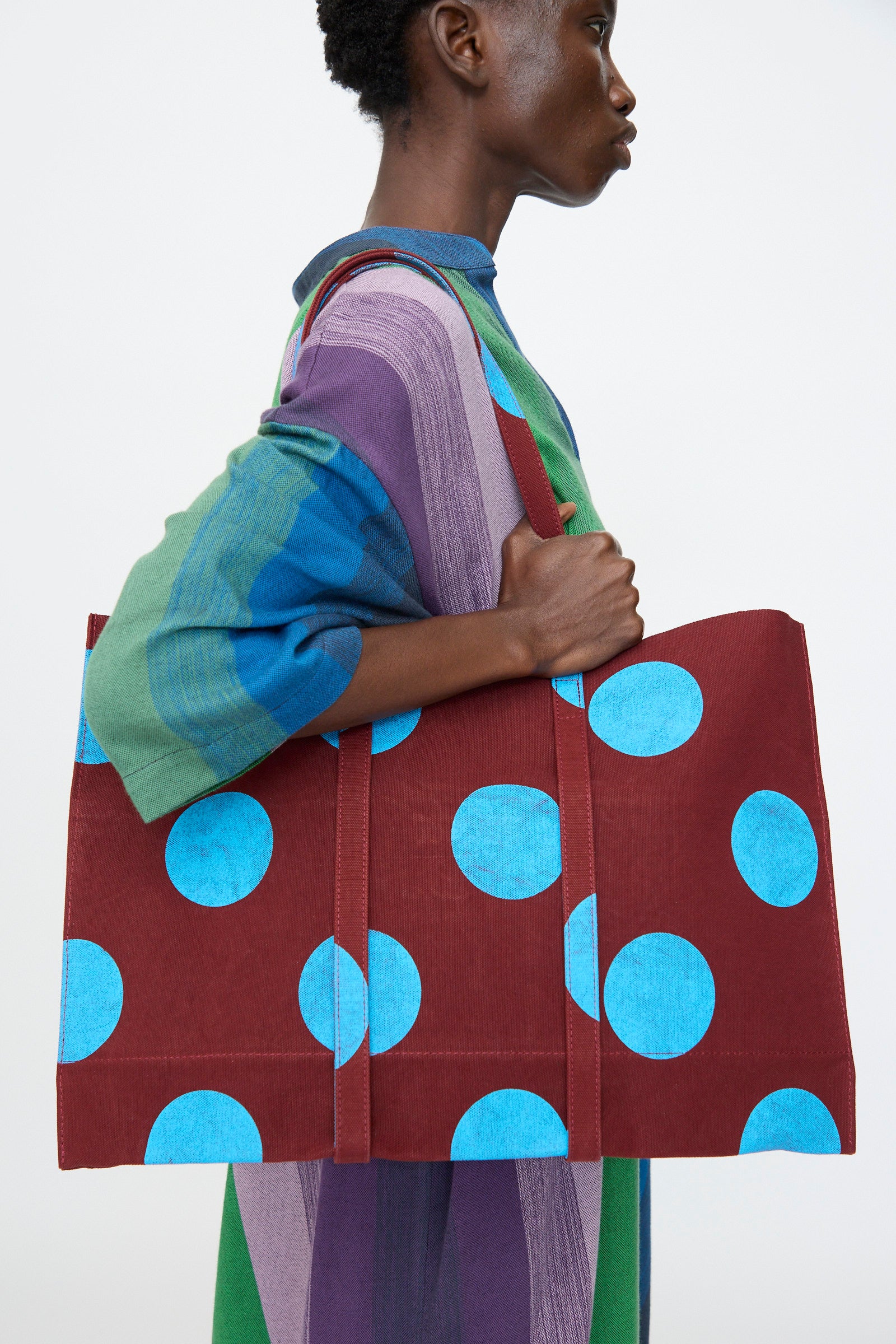 A person carries the Amiacalva Washed Canvas Medium Dot Tote in Sky Blue over their shoulder, adding a playful touch to their striped multicolored dress against a plain background.
