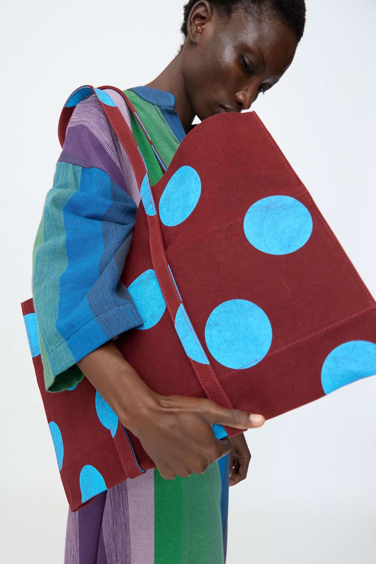 A person in a colorful striped outfit holds Amiacalva’s Washed Canvas Medium Dot Tote in Sky Blue—made in Japan, featuring bright blue dots on a large maroon canvas bag—set against a plain background.