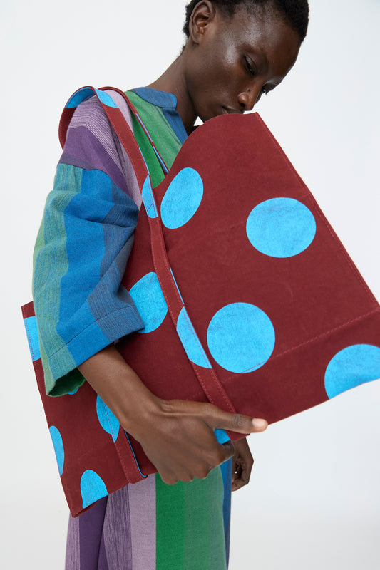 A person in a colorful striped outfit holds Amiacalva’s Washed Canvas Medium Dot Tote in Sky Blue—made in Japan, featuring bright blue dots on a large maroon canvas bag—set against a plain background.