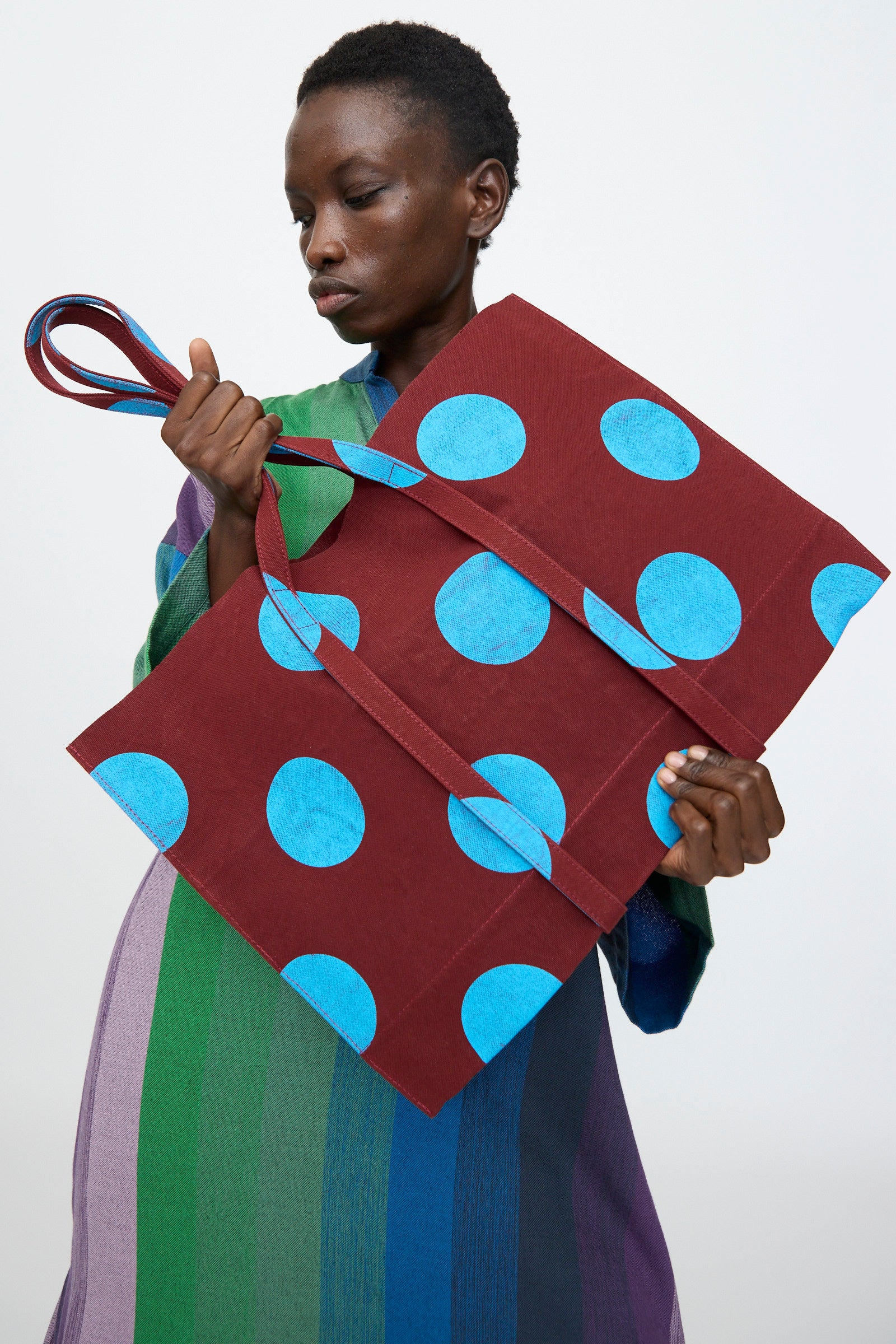 A person holds the Amiacalva Washed Canvas Medium Dot Tote in Sky Blue, made in Japan, while wearing a long striped dress in green, purple, and blue against a plain background.
