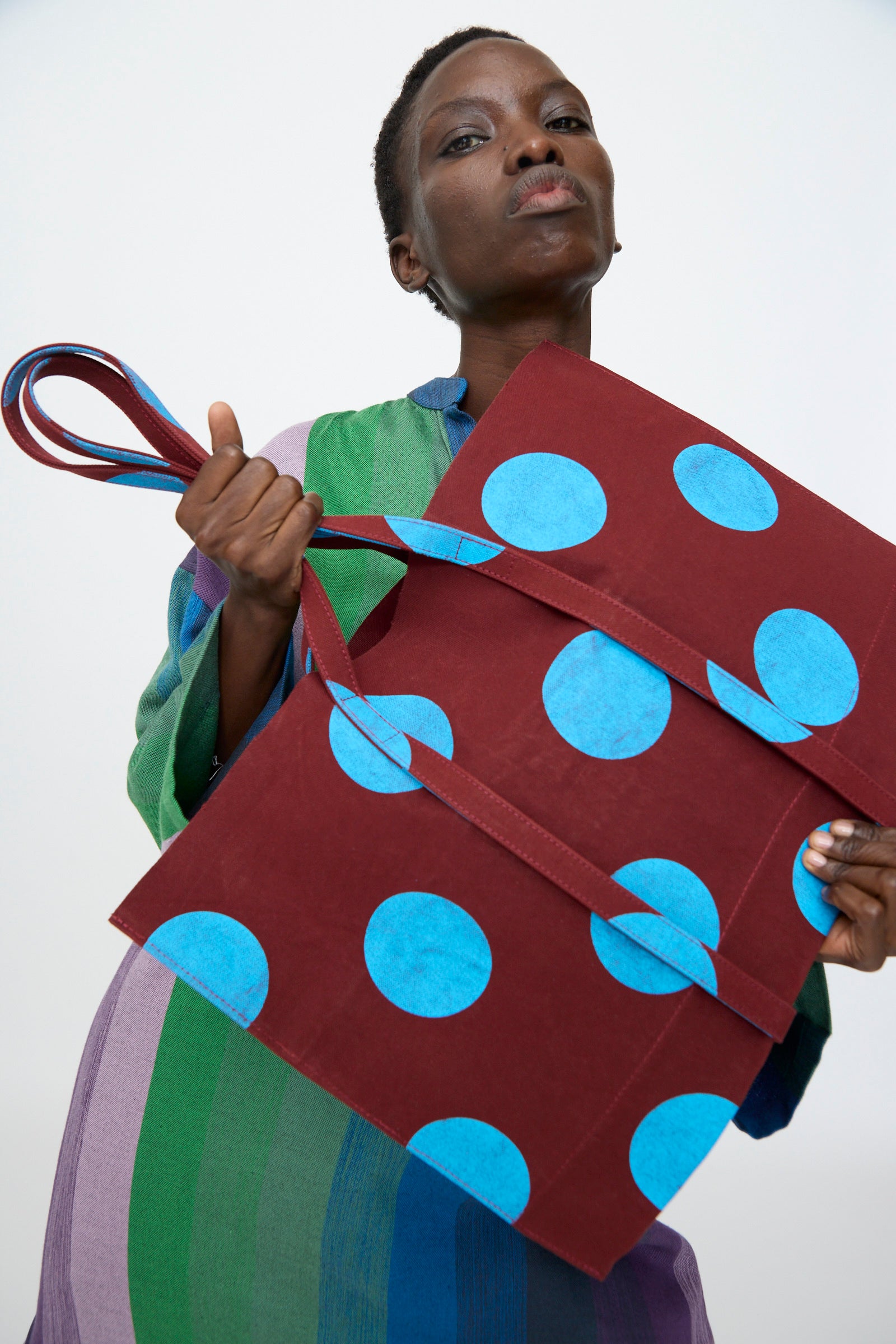 A person in a multicolored striped outfit holds the Amiacalva Washed Canvas Medium Dot Tote in Sky Blue—this chic Japanese-made tote features blue polka dots and matching handles, standing out against a plain background.