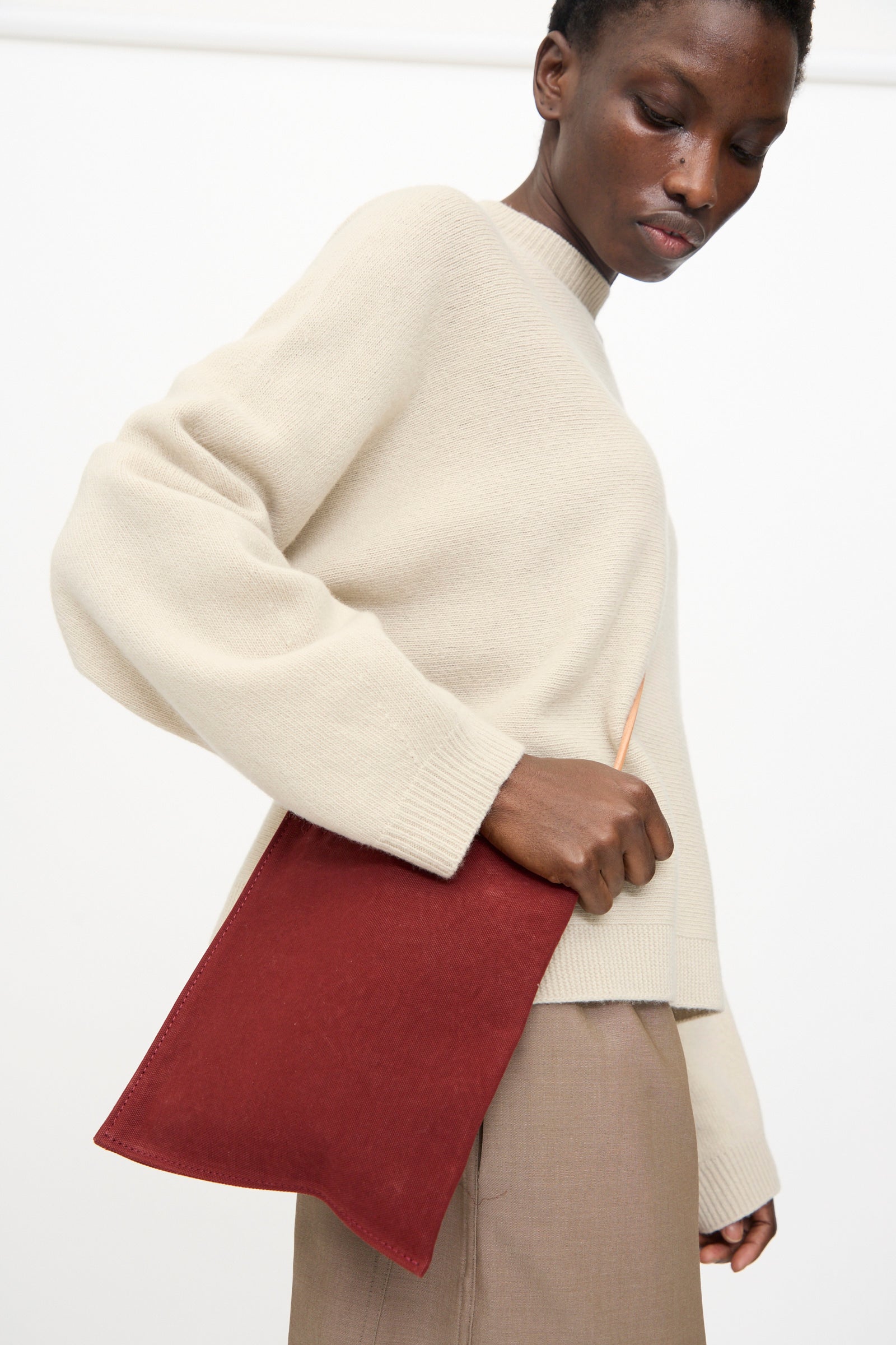 A person in a beige sweater and taupe pants holds the Amiacalva Washed Canvas Pochette in Burgundy—a flat, rectangular bag—by their side.