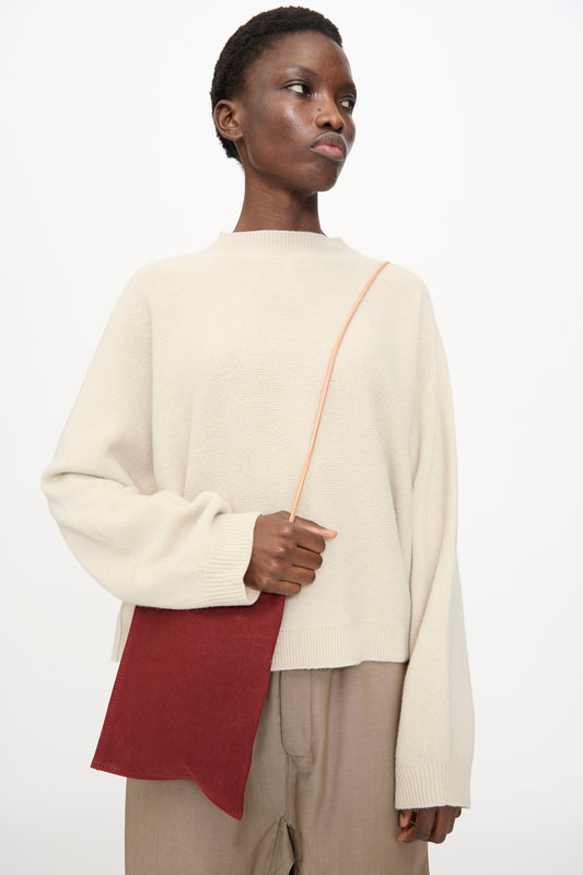 A person wearing a cream sweater and beige pants holds the Amiacalva Washed Canvas Pochette in Burgundy with a tan strap across their body, standing against a plain white background.