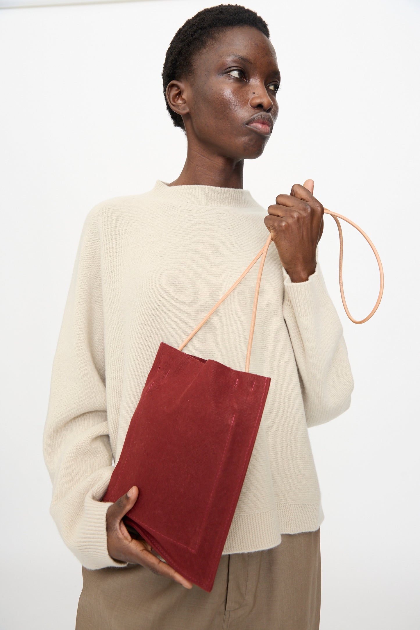 A person wears a beige sweater and tan pants, holding the Amiacalva Washed Canvas Pochette in Burgundy. The rectangular bag features a tan strap and is set against a plain white background.