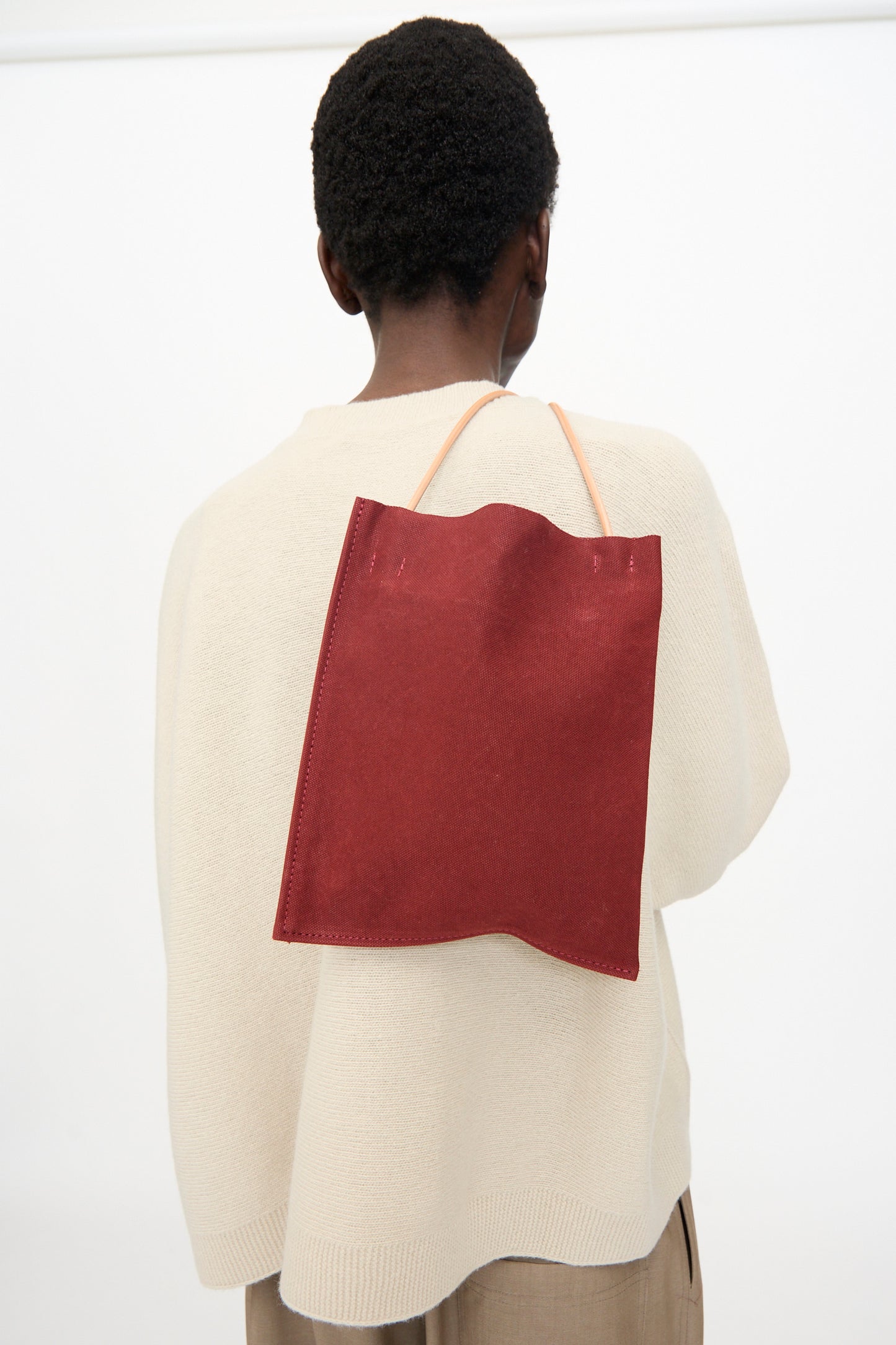Seen from behind, a person in a cream sweater and brown pants carries the Amiacalva Washed Canvas Pochette in Burgundy, a flat, rectangular bag with thin straps, crafted from washed canvas and made in Japan.