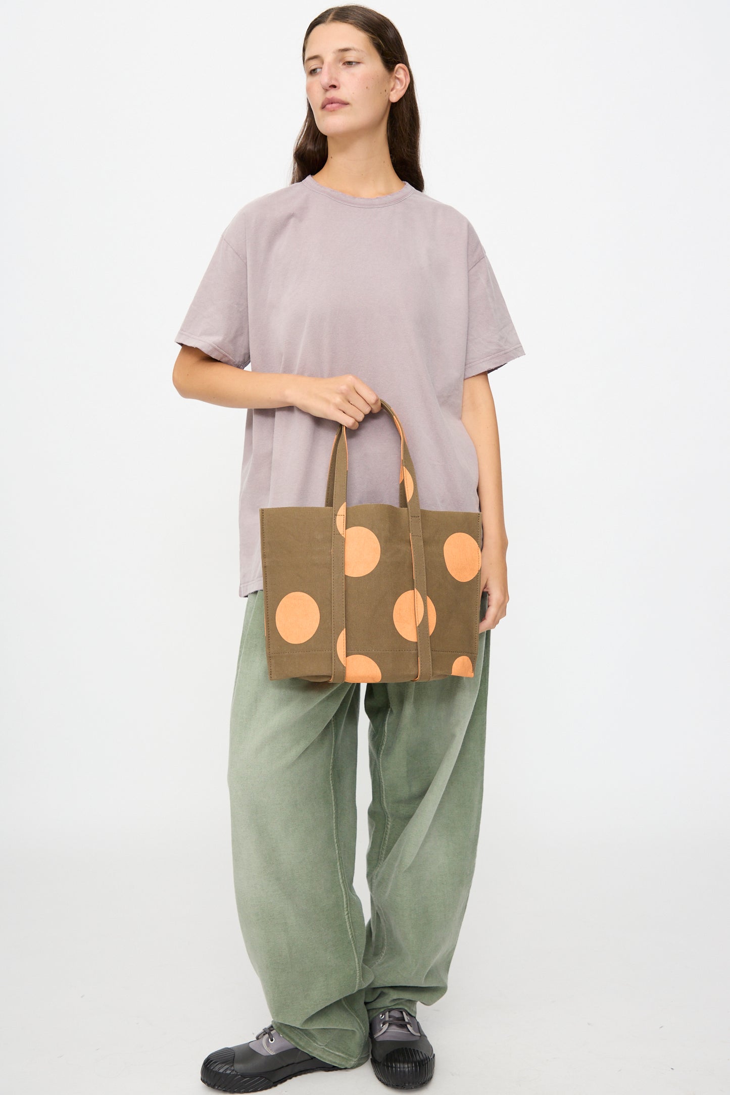 A person stands holding the Amiacalva Washed Canvas Small Dot Tote in Orange—crafted in Japan and featuring bold orange polka dots—paired with a loose lavender t-shirt, green wide-leg pants, and black shoes against a plain white background.