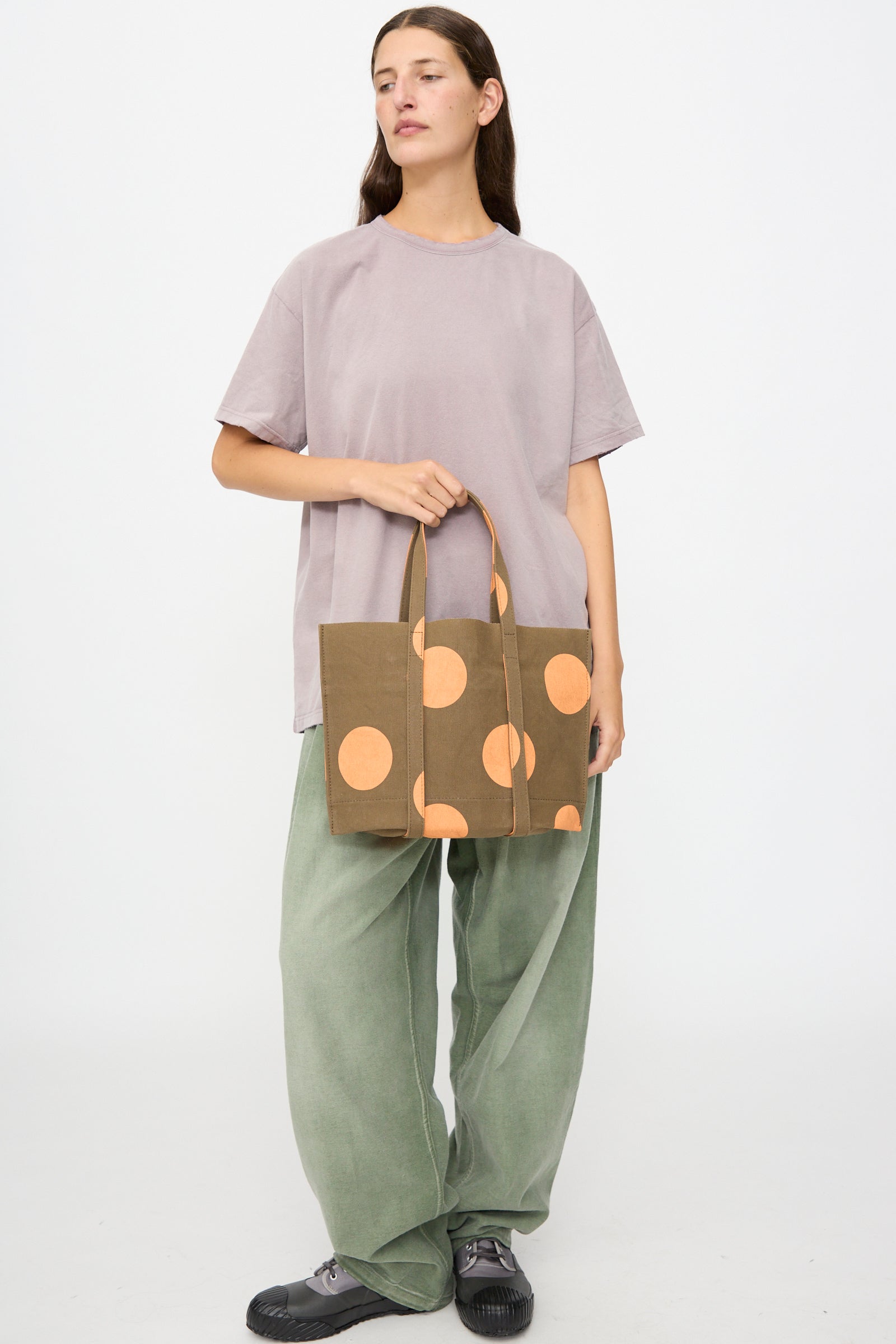 A person stands holding the Amiacalva Washed Canvas Small Dot Tote in Orange—crafted in Japan and featuring bold orange polka dots—paired with a loose lavender t-shirt, green wide-leg pants, and black shoes against a plain white background.