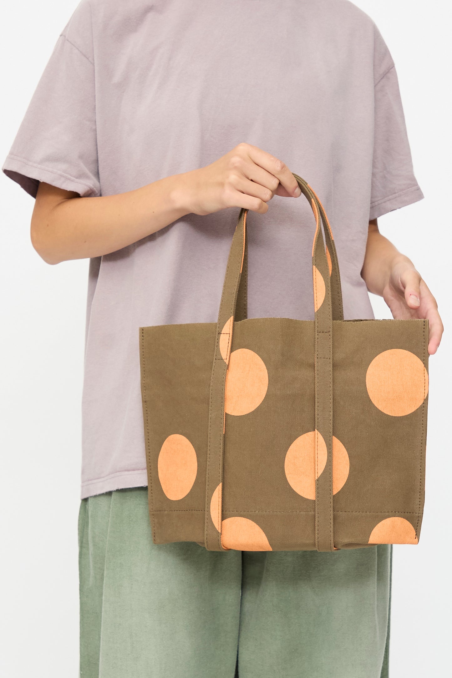 A person holds the Amiacalva Washed Canvas Small Dot Tote in Orange—a khaki canvas bag with large orange polka dots, made in Japan—while wearing a light purple shirt and green pants against a plain background.