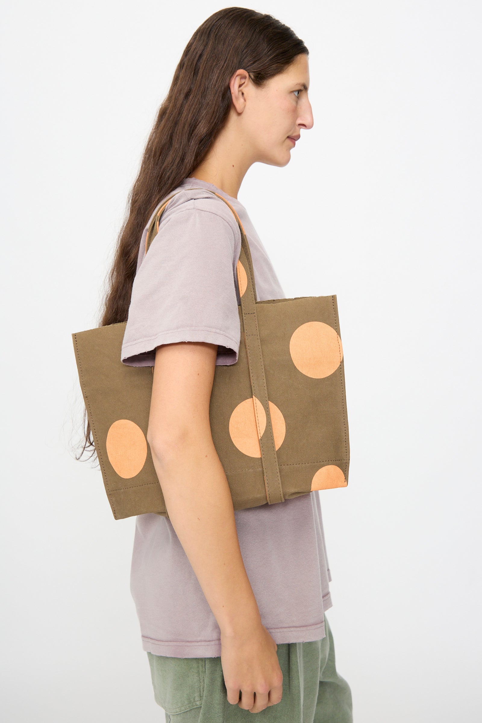 A person in a light purple shirt carries the Amiacalva Washed Canvas Small Dot Tote in Orange, made in Japan, as they stand against a plain white background.