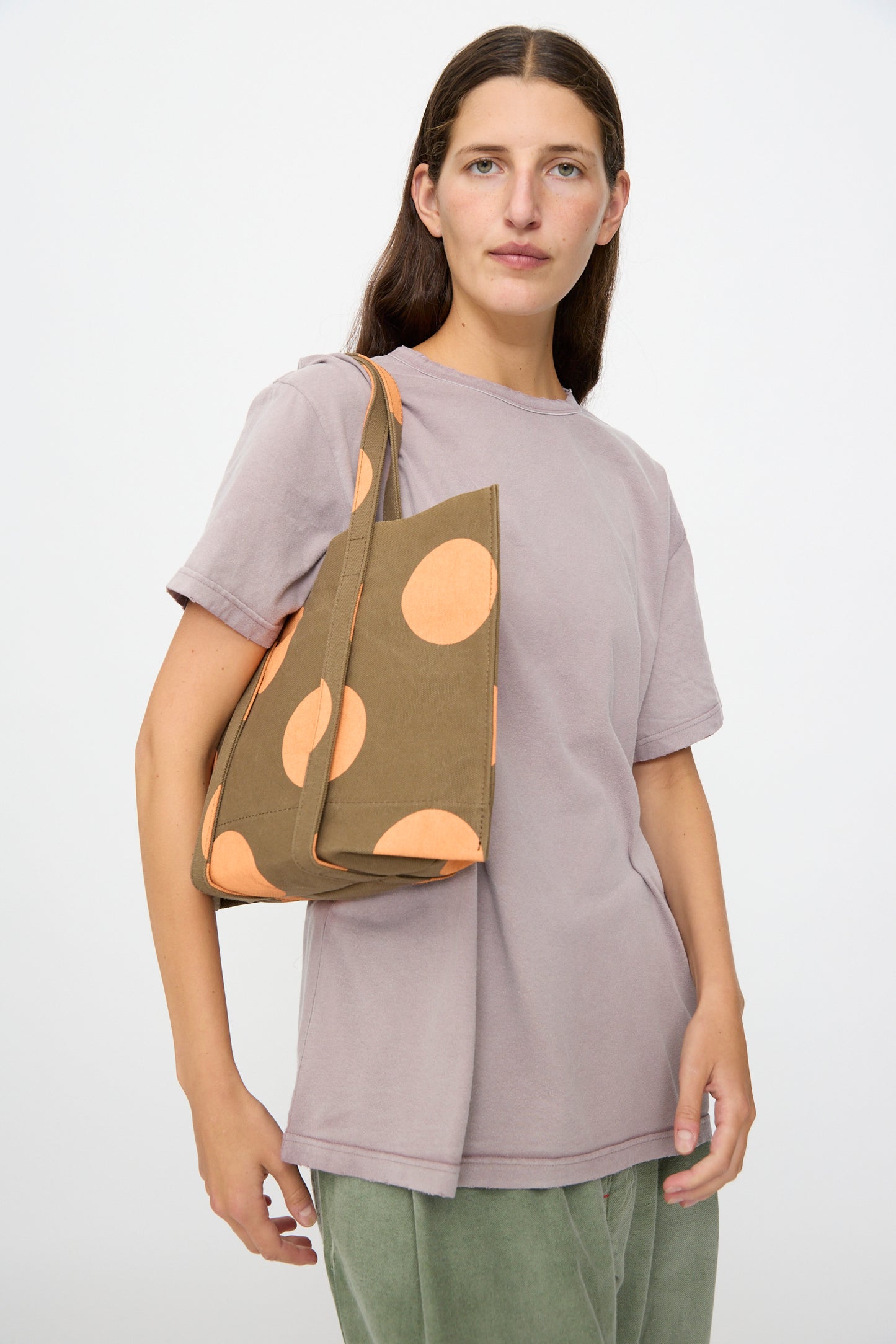 A woman with long brown hair, dressed in a lavender t-shirt and green pants, carries the Amiacalva Washed Canvas Small Dot Tote in Orange—crafted in Japan—as she stands against a plain background.