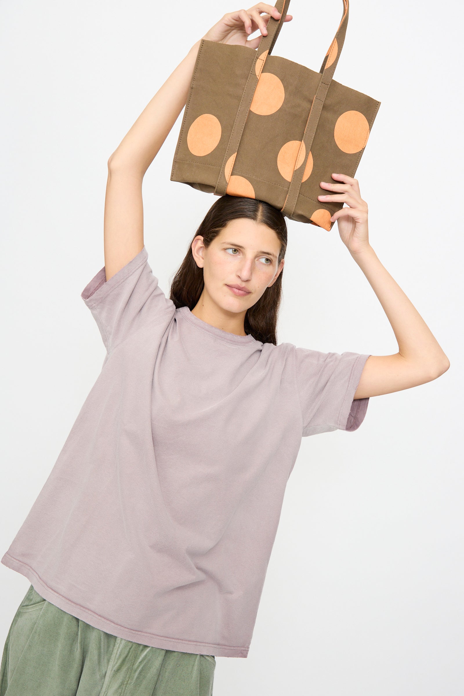 A woman in a loose lavender t-shirt holds the Amiacalva Washed Canvas Small Dot Tote in Orange above her head against a plain white background.