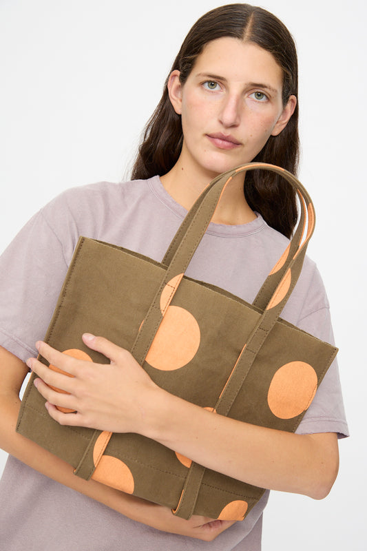 A woman with long brown hair, dressed in a light purple t-shirt, stands against a plain white background holding the Amiacalva Washed Canvas Small Dot Tote in Orange—an olive green tote bag with large orange polka dots made in Japan.