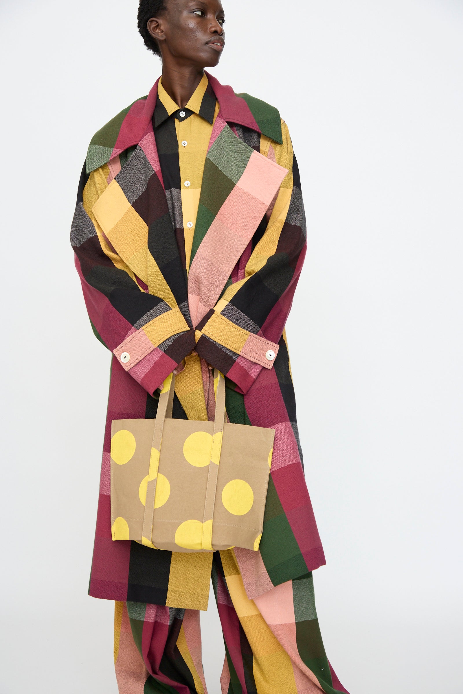 A person in a multicolored plaid coat and matching pants holds the Amiacalva Washed Canvas Small Dot Tote in Yellow, made in Japan, standing against a plain background.