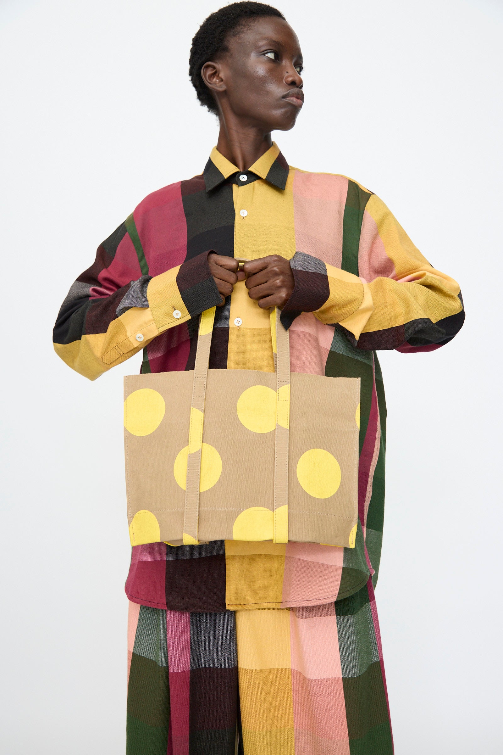 A person wearing a colorful, oversized plaid outfit holds the Amiacalva Washed Canvas Small Dot Tote in Yellow against a plain white background.