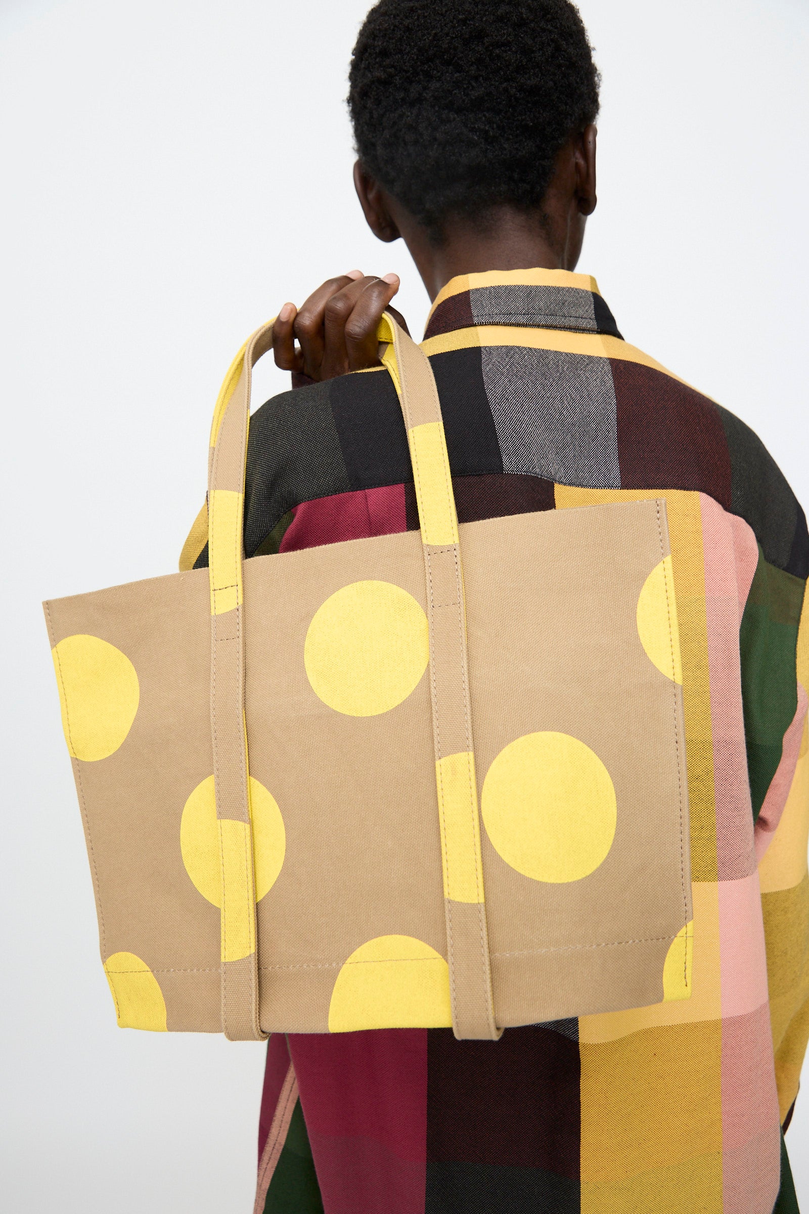 Person facing away, wearing a multicolored plaid shirt and carrying Amiacalva’s Washed Canvas Small Dot Tote in Yellow over their shoulder. This stylish Japanese tote combines playful design with everyday practicality.