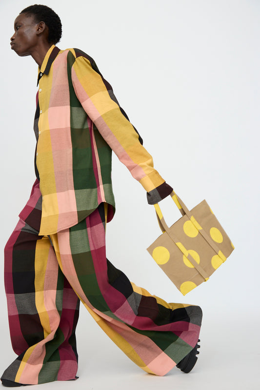 A person walks against a plain white background, wearing an oversized multicolored plaid shirt and pants, holding the Amiacalva Washed Canvas Small Dot Tote in Yellow.