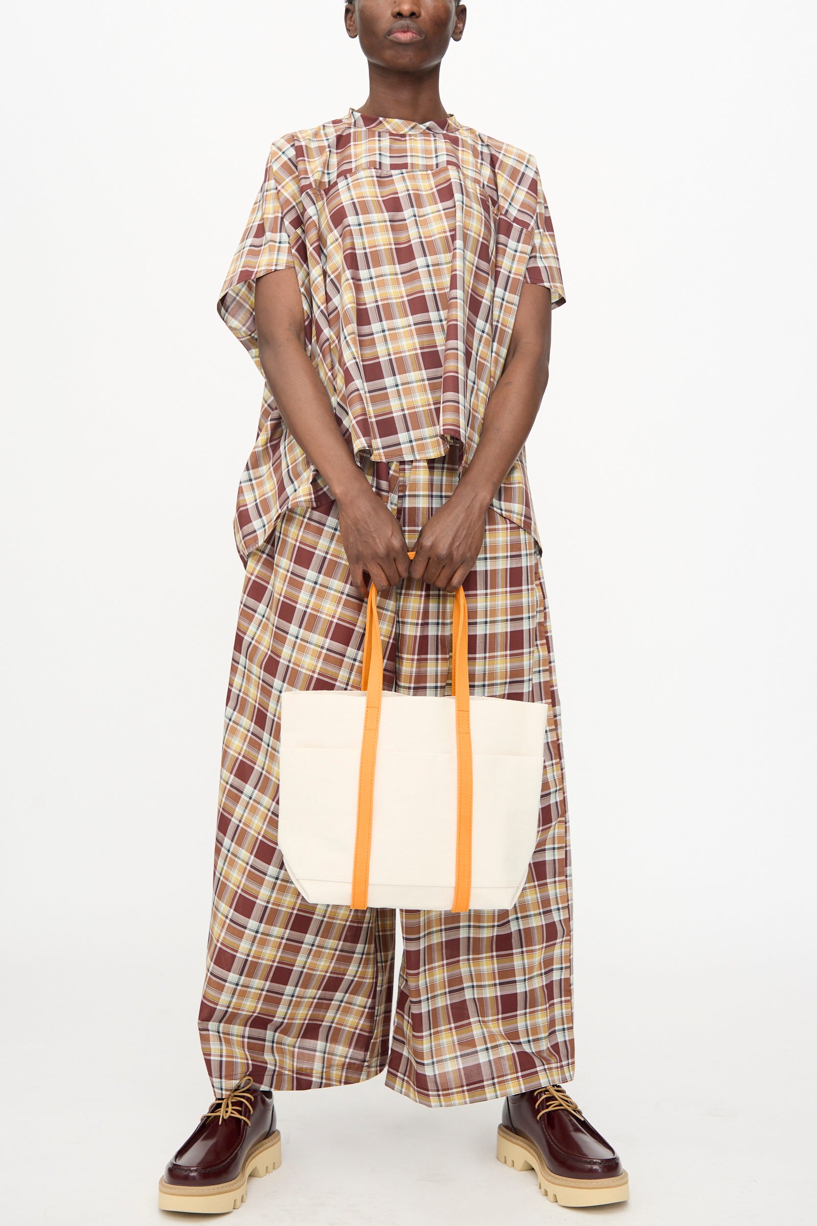 A person holds the Amiacalva Washed Canvas Small Tote 2 Tone in Orange, made in Japan; they wear a loose plaid outfit and brown thick-soled shoes.