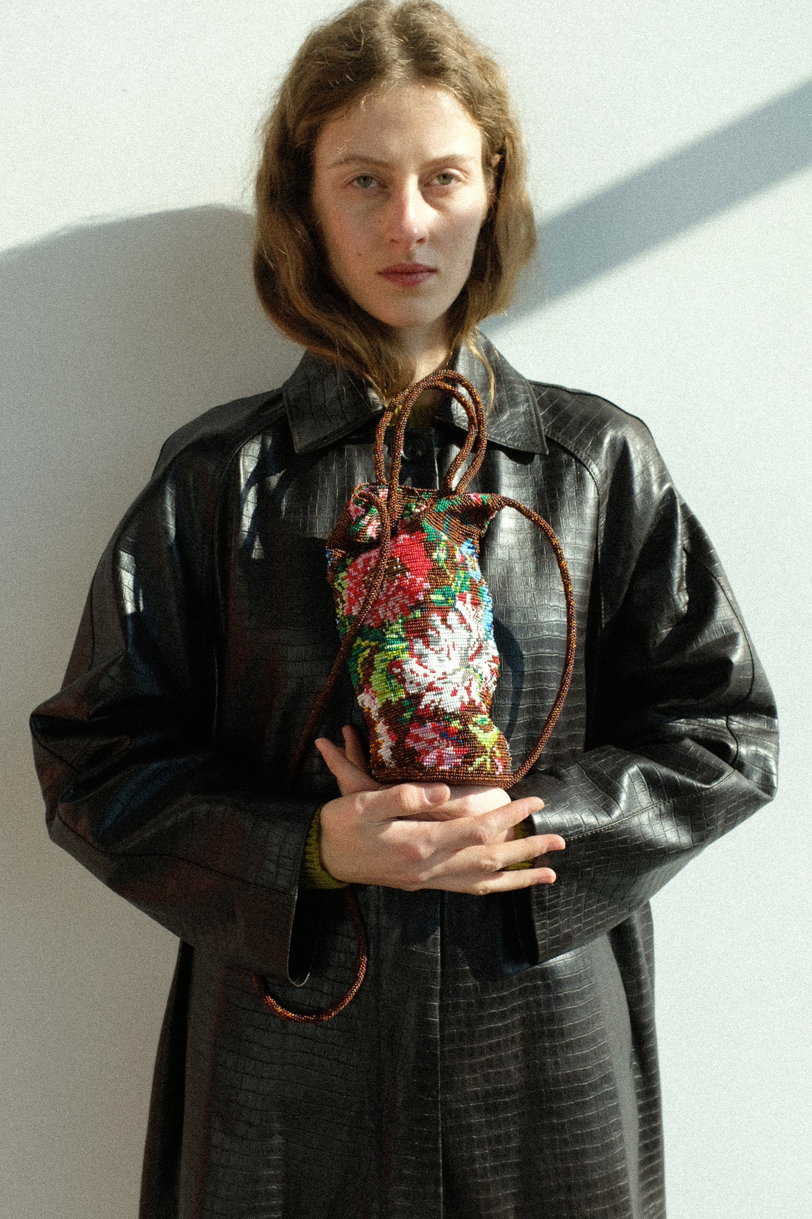 A woman holding Cordera's Handmade Beaded Bag in Peonia, wearing the Nappa Long Coat in Crocodile against a white wall.