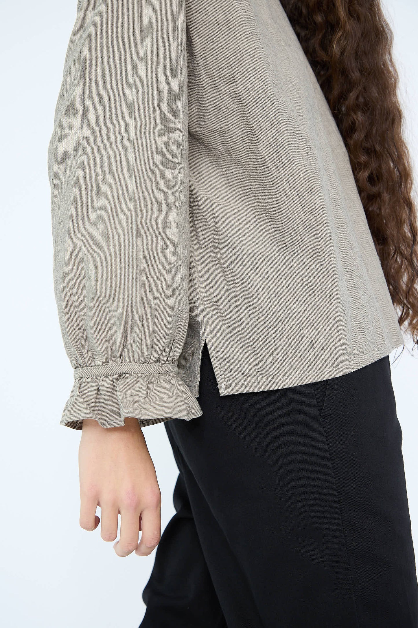 A person wearing an Antiquités Noir Cotton Linen Two Way Frill Blouse in Stripe and black pants. Their arm is relaxed at their side, and the background is plain white.