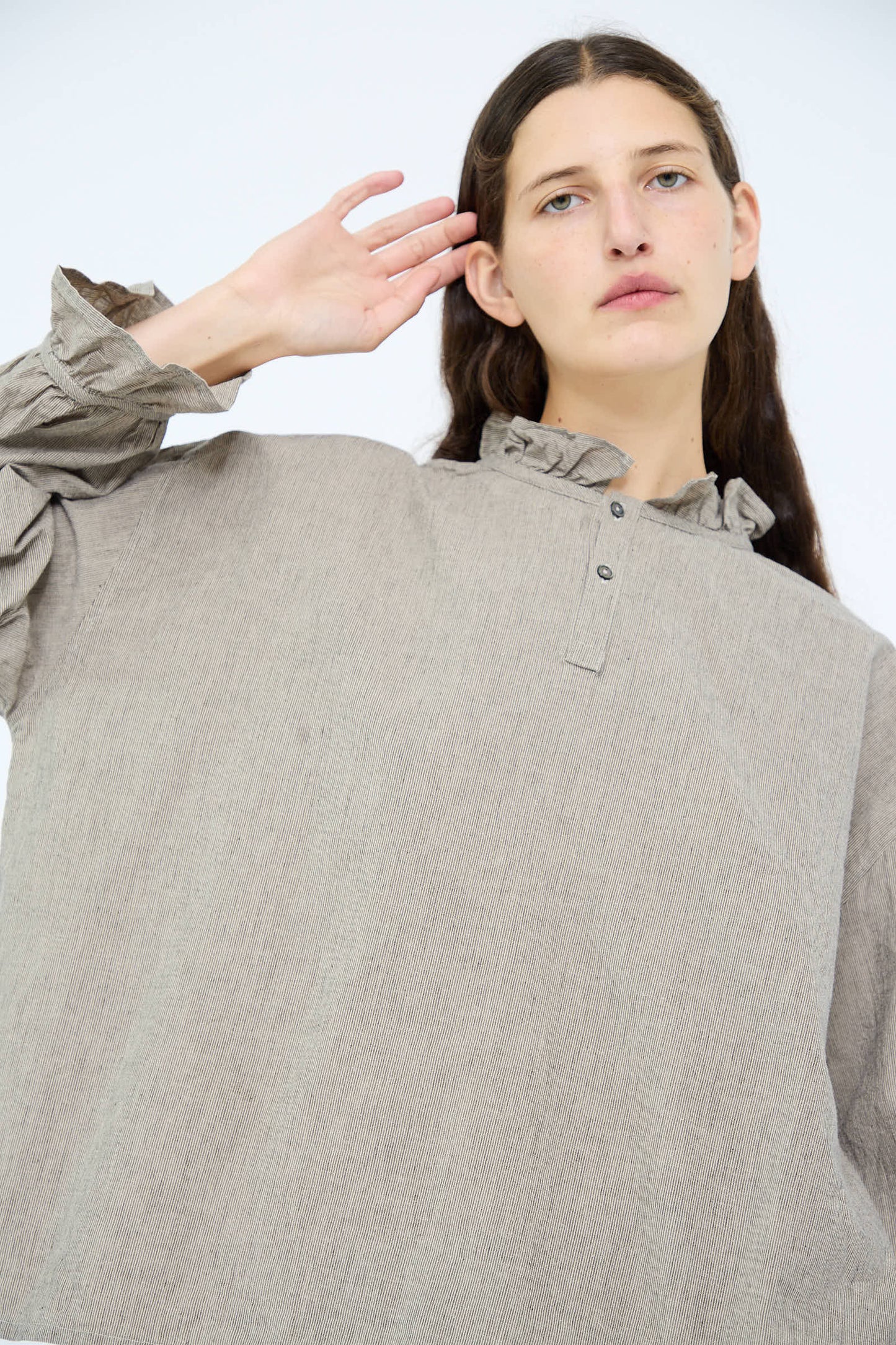 A person wearing the Antiquités Noir Cotton Linen Two Way Frill Blouse in Stripe, featuring ruffle detail on the collar and cuffs, holds one hand near their ear.