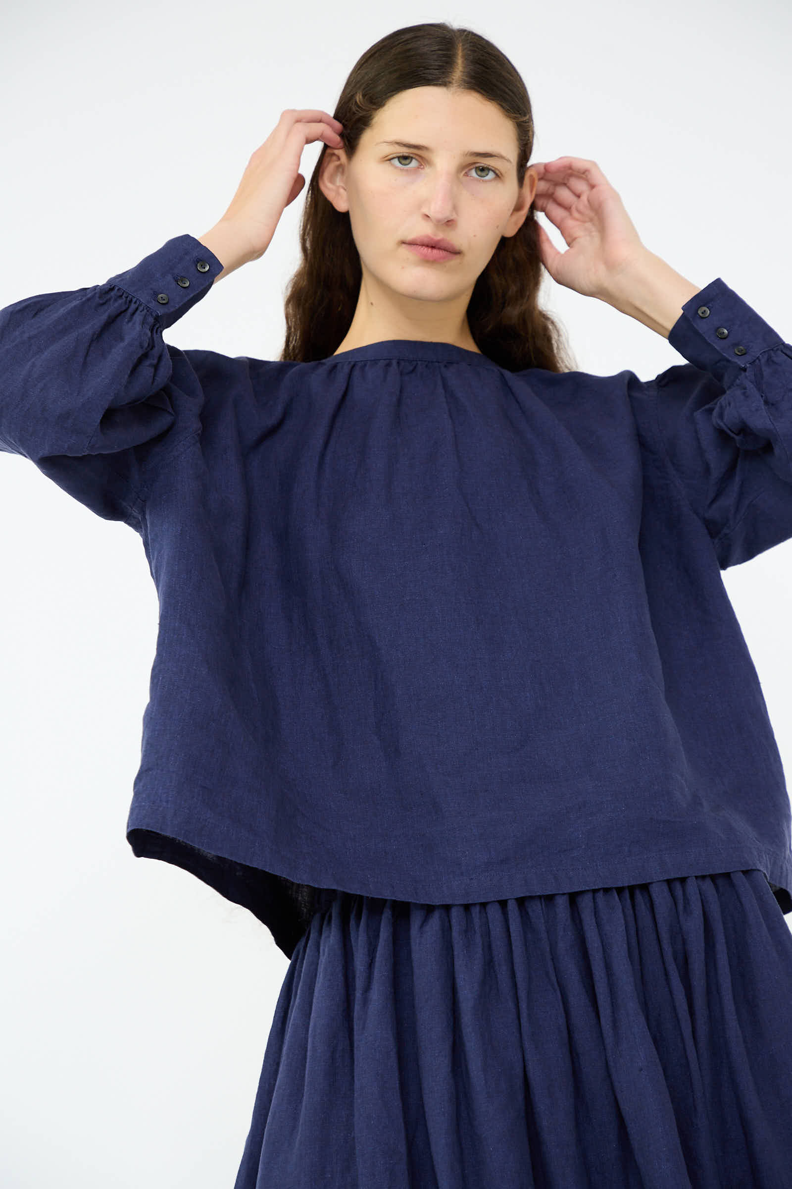 A person wearing the Linen Band Collar Blouse in Indigo by Antiquités Noir, along with a matching skirt featuring a button back closure, is adjusting their hair against a plain backdrop.