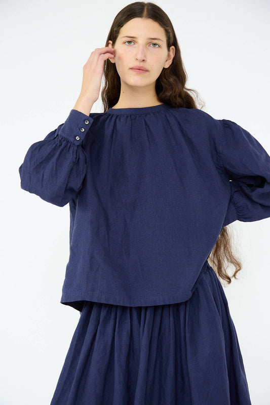 A person with long hair wearing the relaxed fit Linen Band Collar Blouse in Indigo by Antiquités Noir stands against a plain background, paired with a matching skirt.