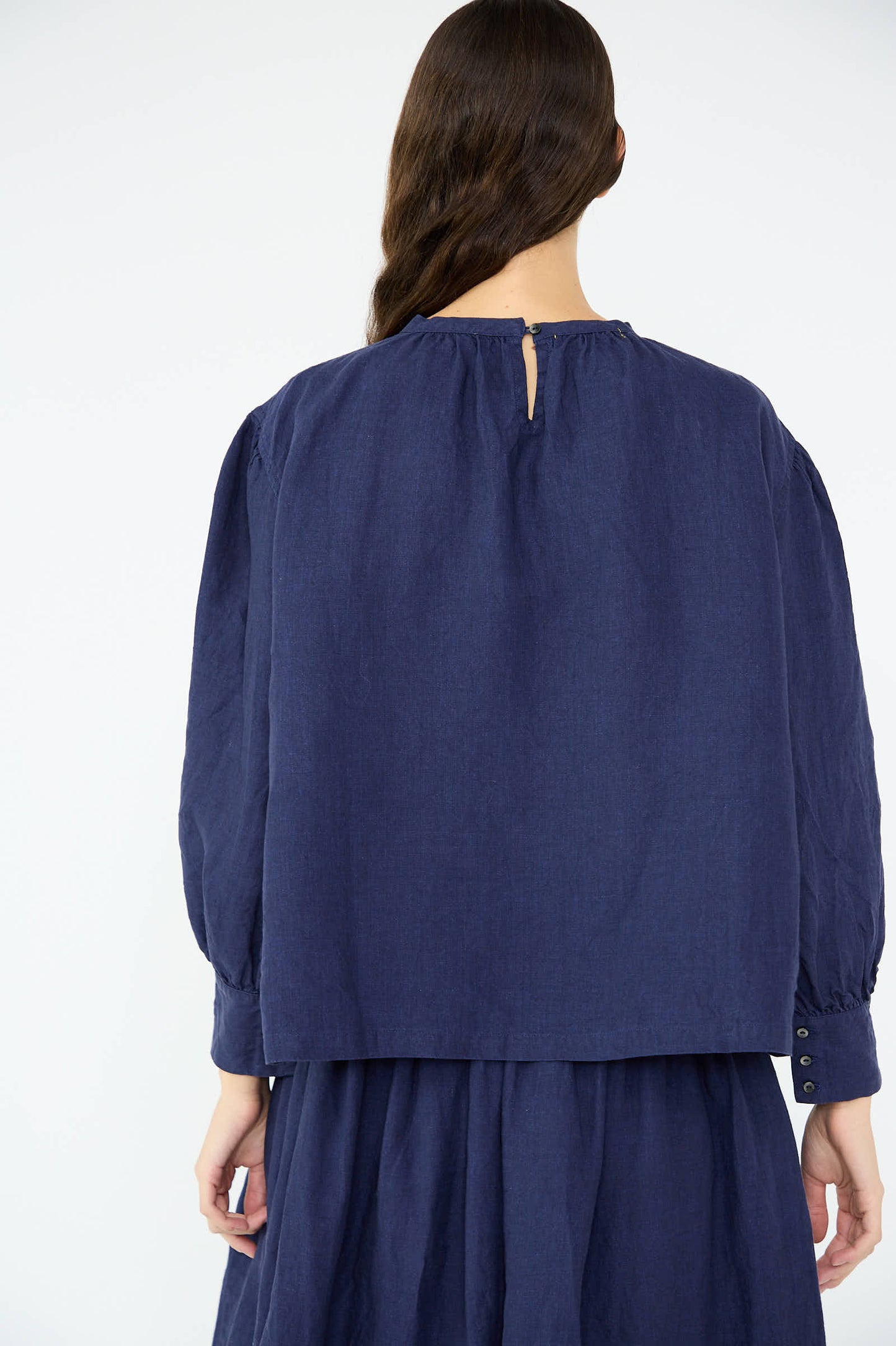 A person with long hair is wearing the Antiquités Noir Linen Band Collar Blouse in Indigo, showcasing its relaxed fit and button-back closure from the back.