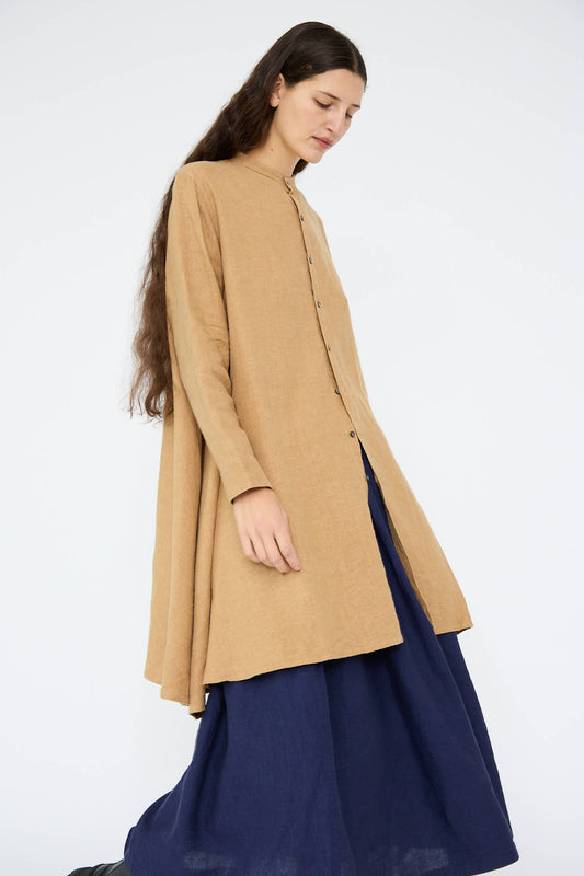 A person with long hair is wearing the Linen Canvas Long Shirt in Camel by Antiquités Noir, paired with navy wide-legged pants, standing against a plain background.