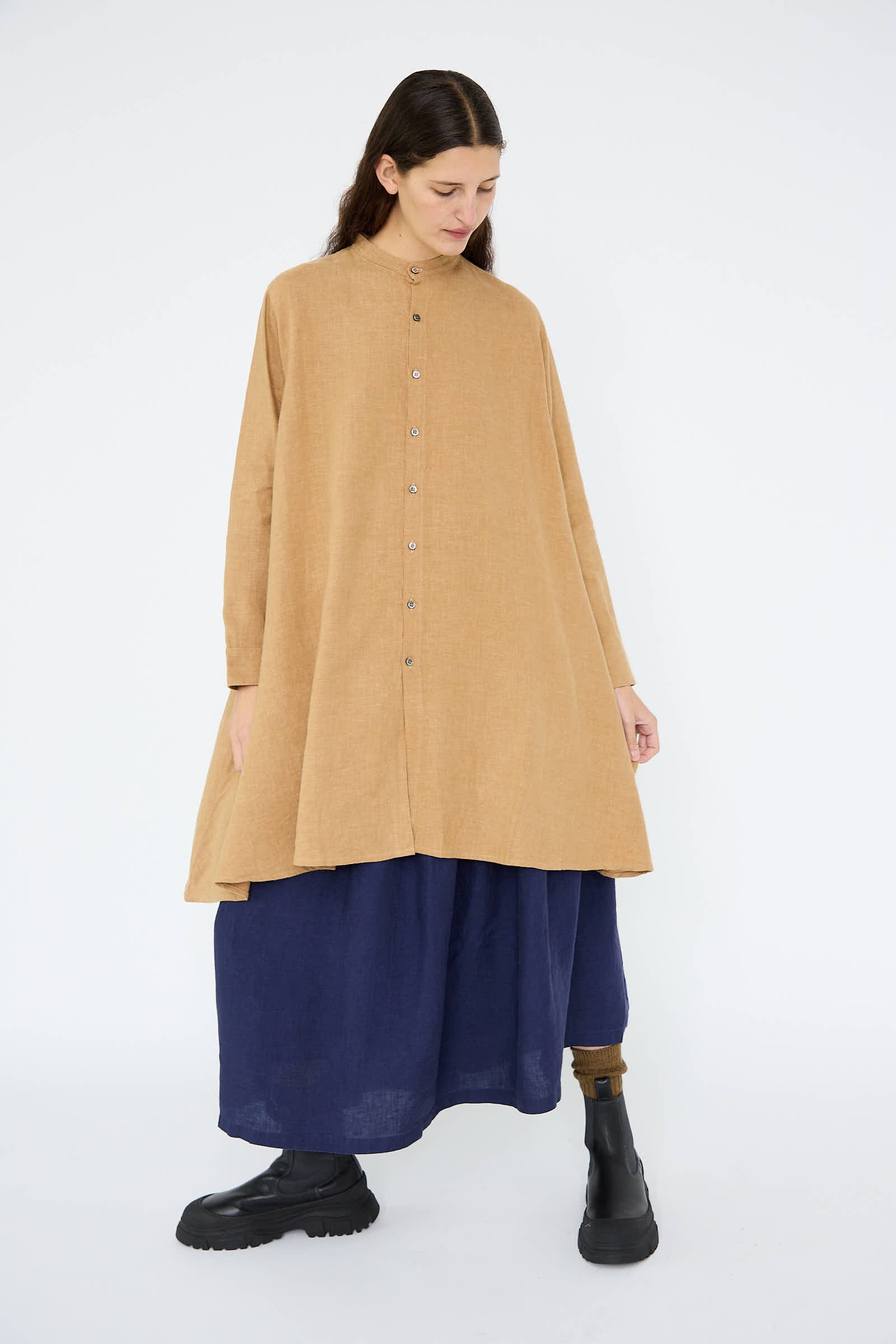 A person wearing the Antiquités Noir Linen Canvas Long Shirt in Camel over a long, dark blue skirt and black boots stands against a plain white background.