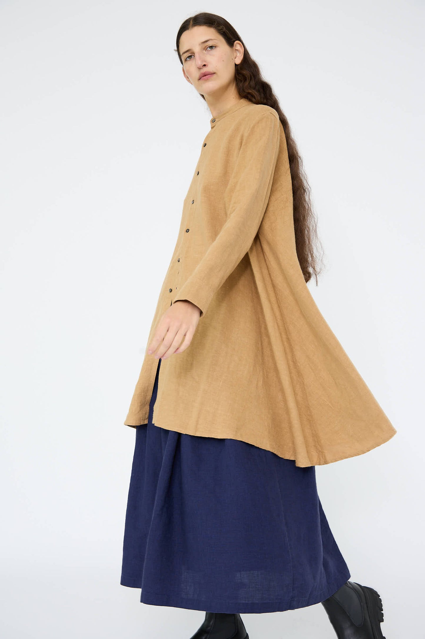 A person with long hair wears a tan, button-up coat and the Linen Canvas Long Shirt in Camel by Antiquités Noir over a dark blue skirt and black shoes, walking against a plain white background.