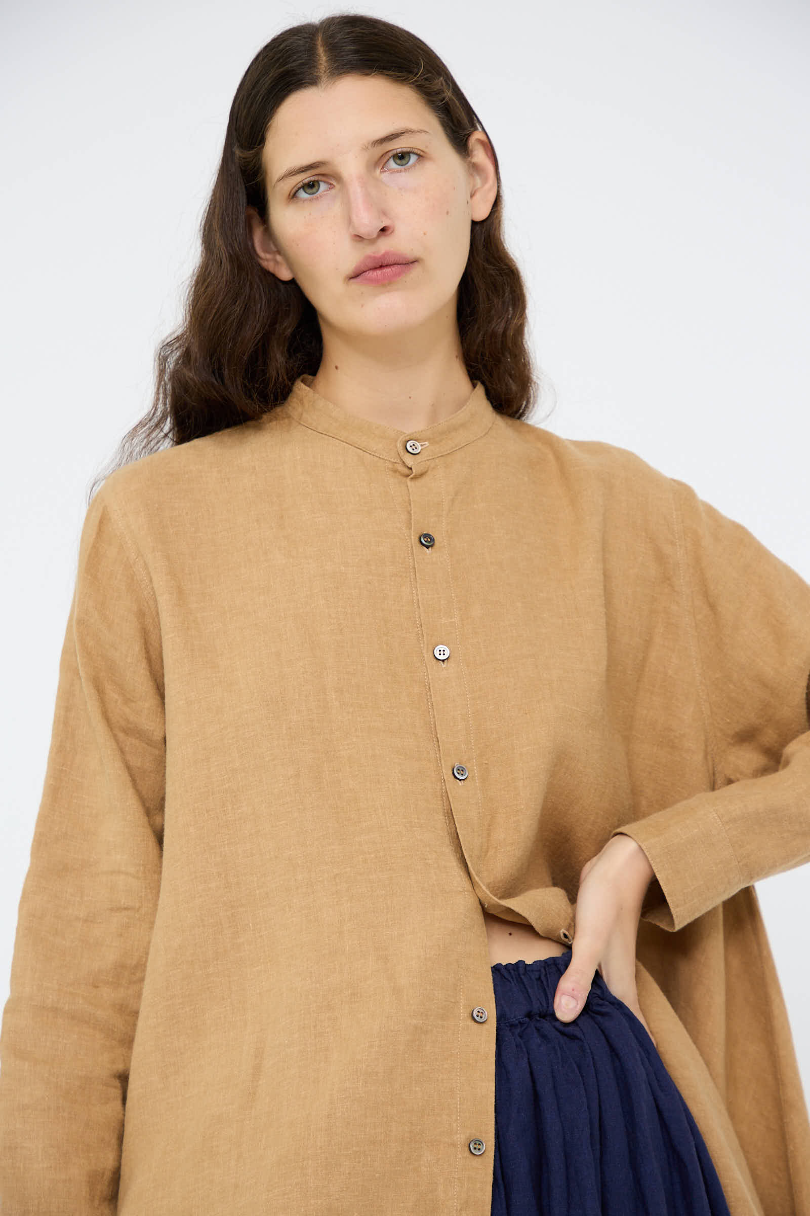 An individual with long dark hair is wearing the Linen Canvas Long Shirt in Camel by Antiquités Noir and dark pants, standing against a plain background.