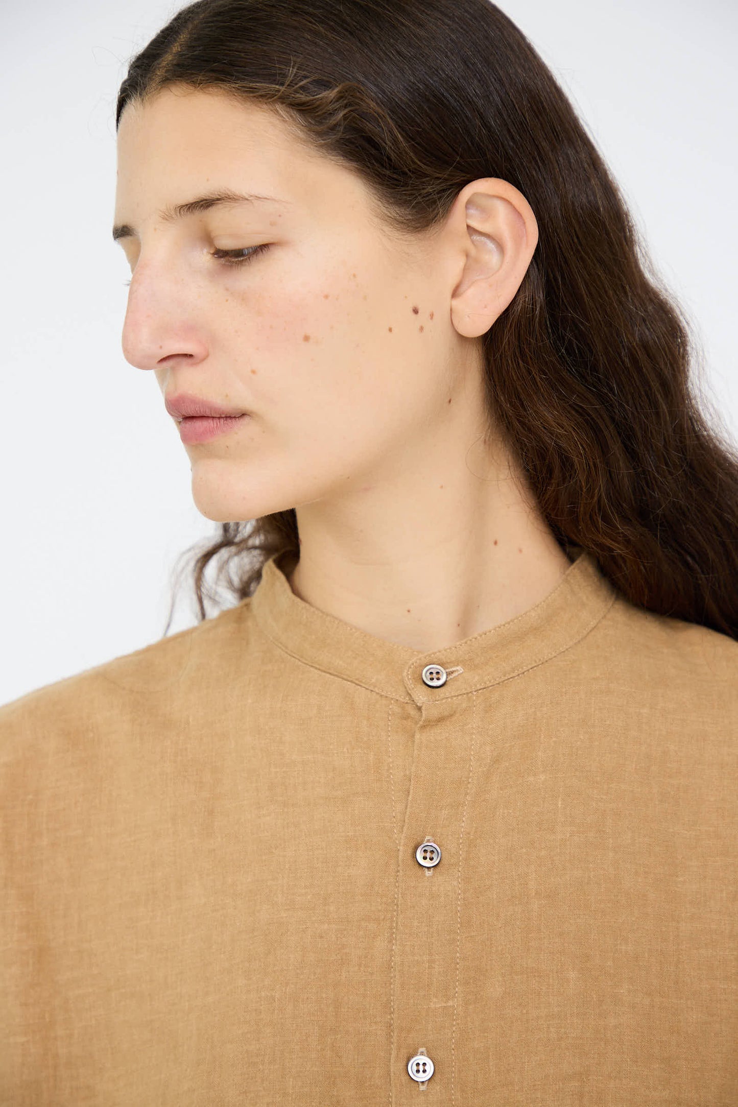 A person with long brown hair, wearing the Antiquités Noir Linen Canvas Long Shirt in Camel, gazes thoughtfully to the side against a plain background.