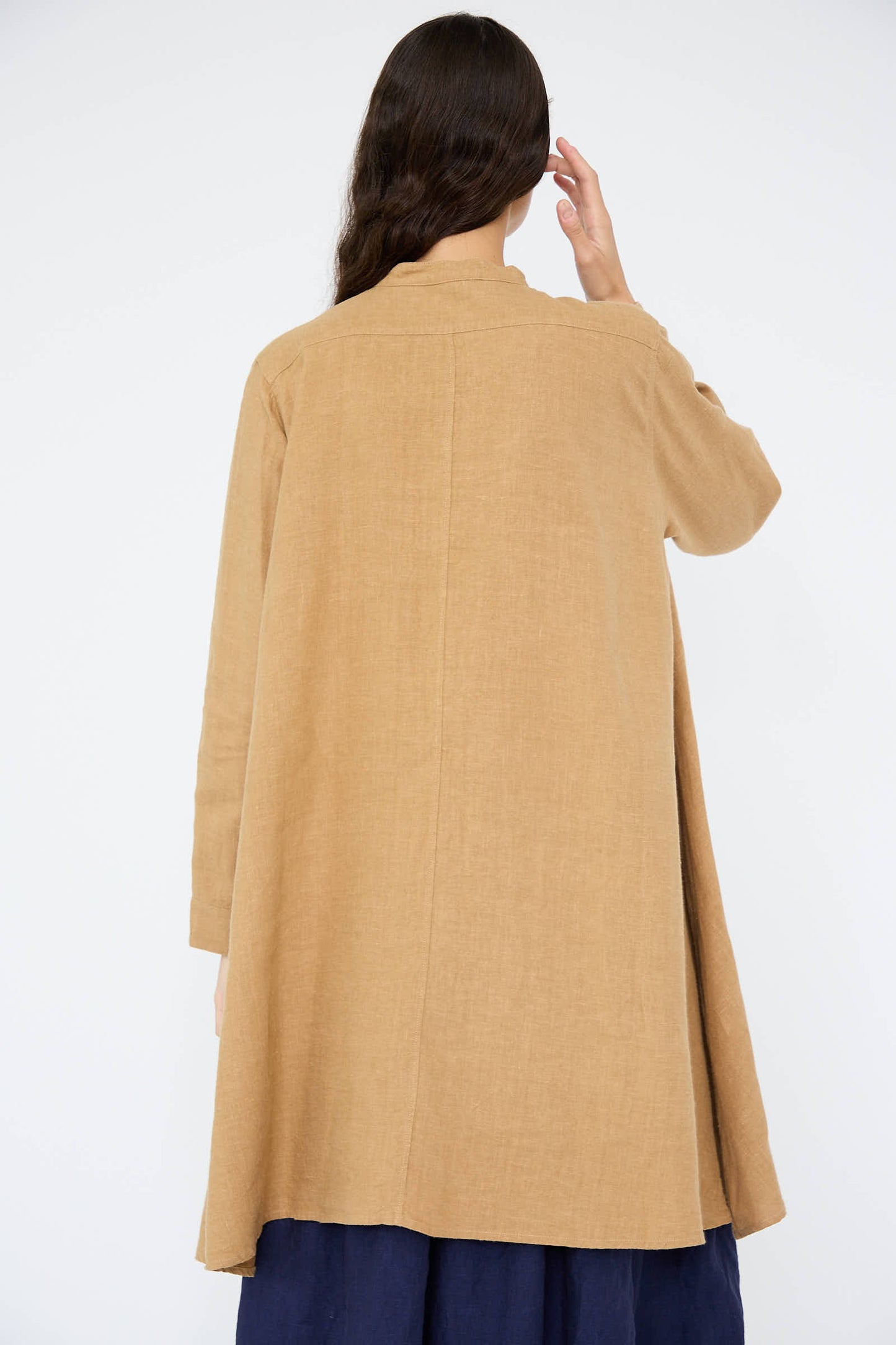 A person with long hair, wrapped in a brown, long-sleeved coat, contrasts against the soft texture of Antiquités Noir's Linen Canvas Long Shirt in Camel peeking through as they face away.