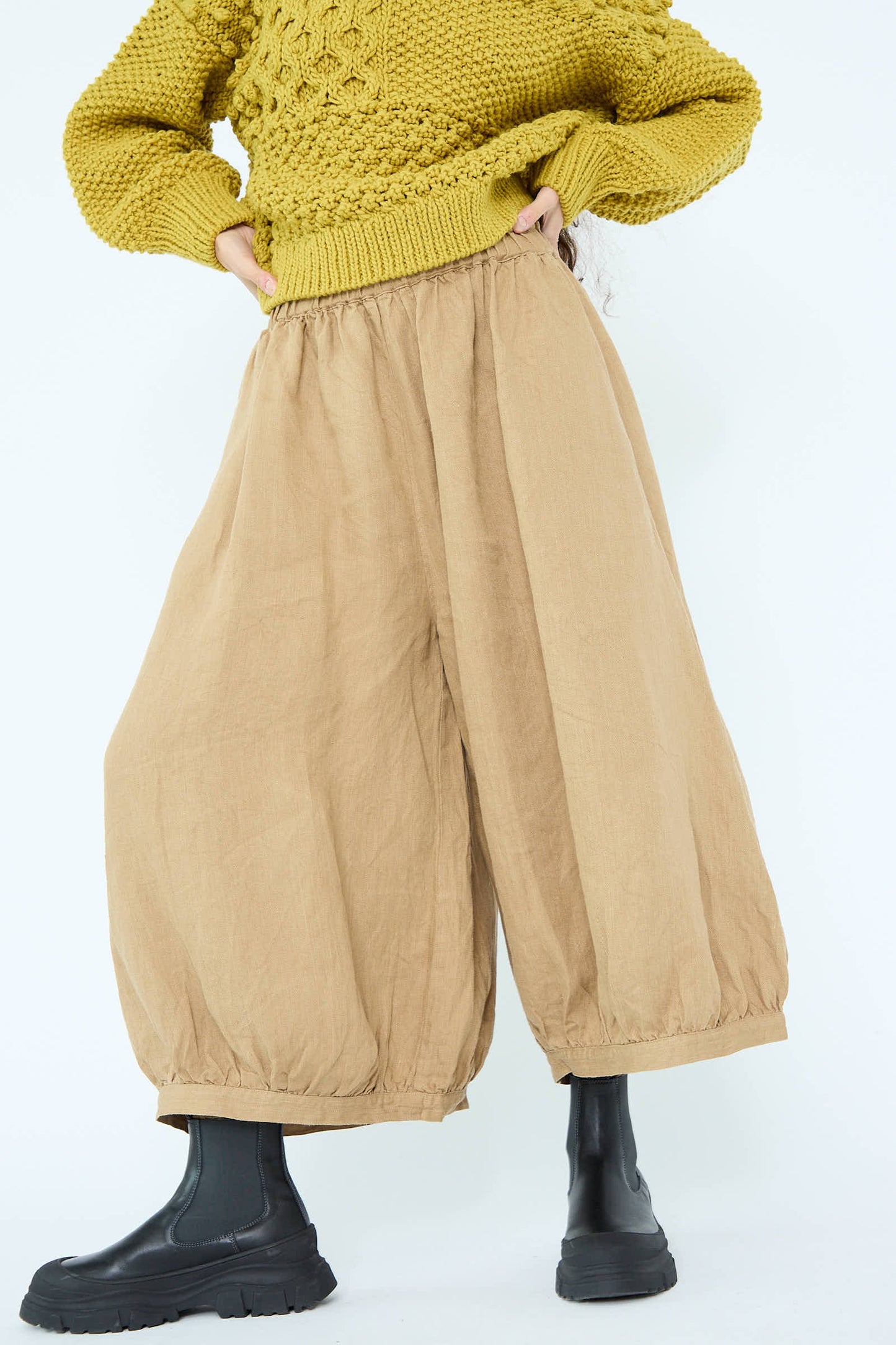 Person wearing a yellow textured sweater, Linen Chino Denim Balloon Pant in Beige by Antiquités Noir with an elasticized waistband, and black boots, standing against a plain background.