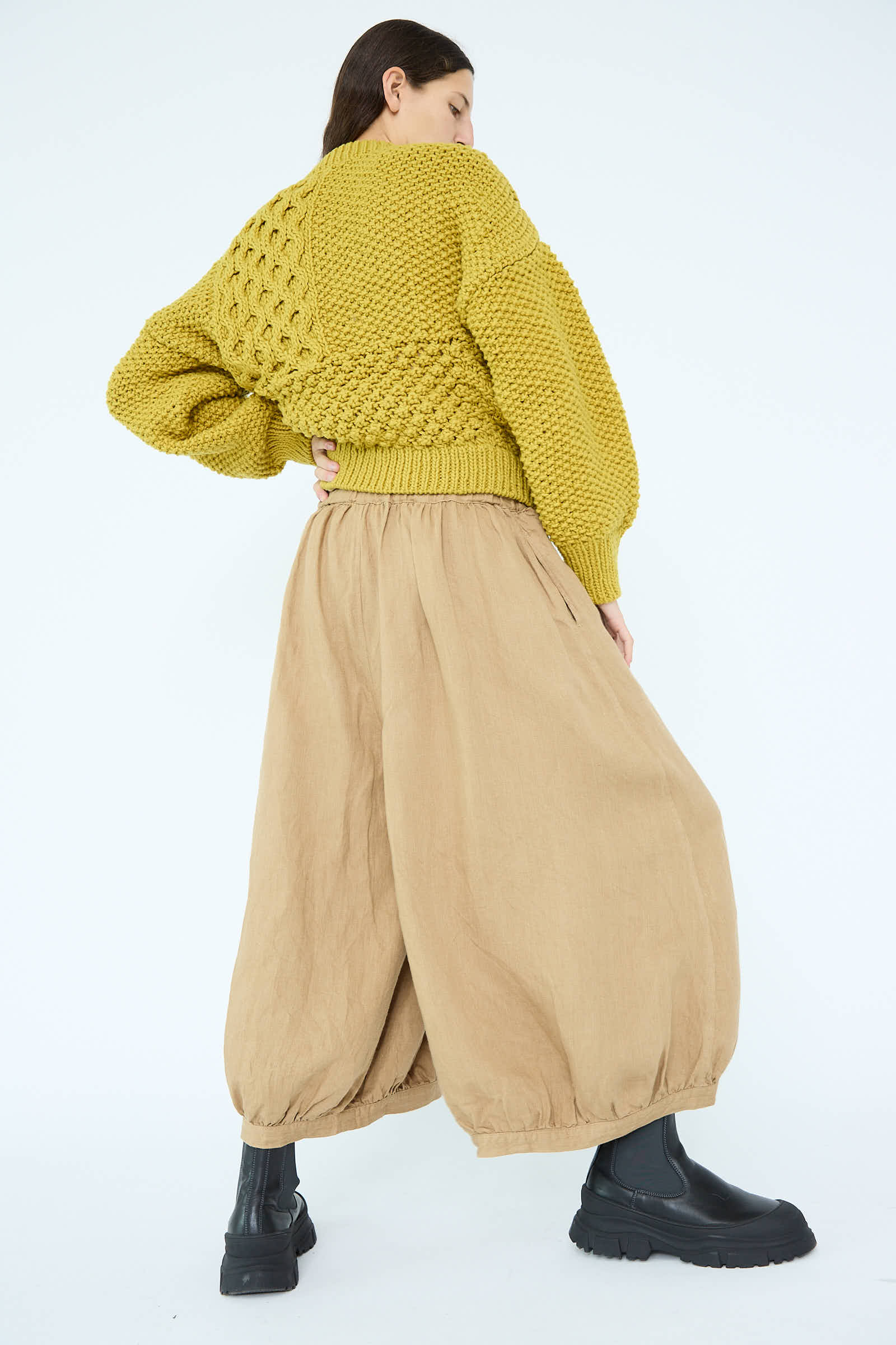 Individual wearing a chunky yellow sweater, Antiquités Noir Linen Chino Denim Balloon Pants in beige with an elasticized waistband, and black boots, posing with their back turned.