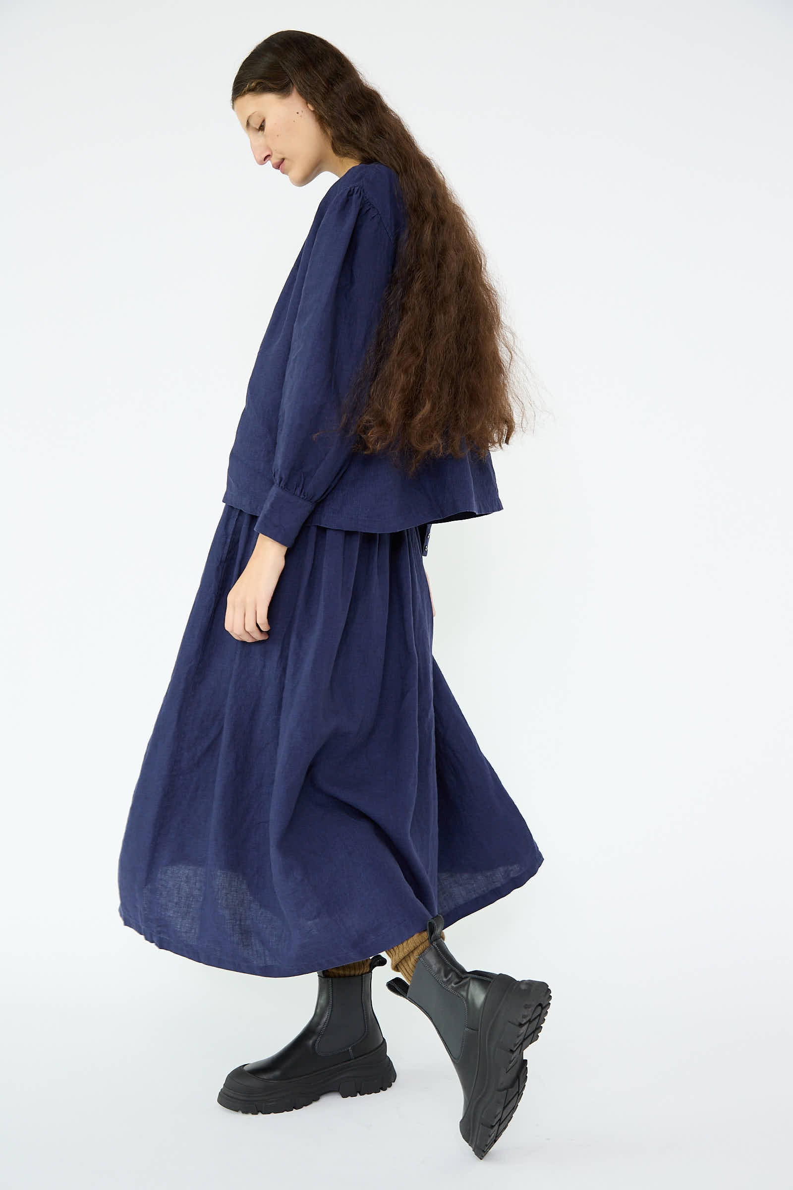 Dressed in the Linen Gather Skirt in Indigo by Antiquités Noir, a person with long hair walks confidently on a white background, their black boots adding a striking contrast to the ensemble.