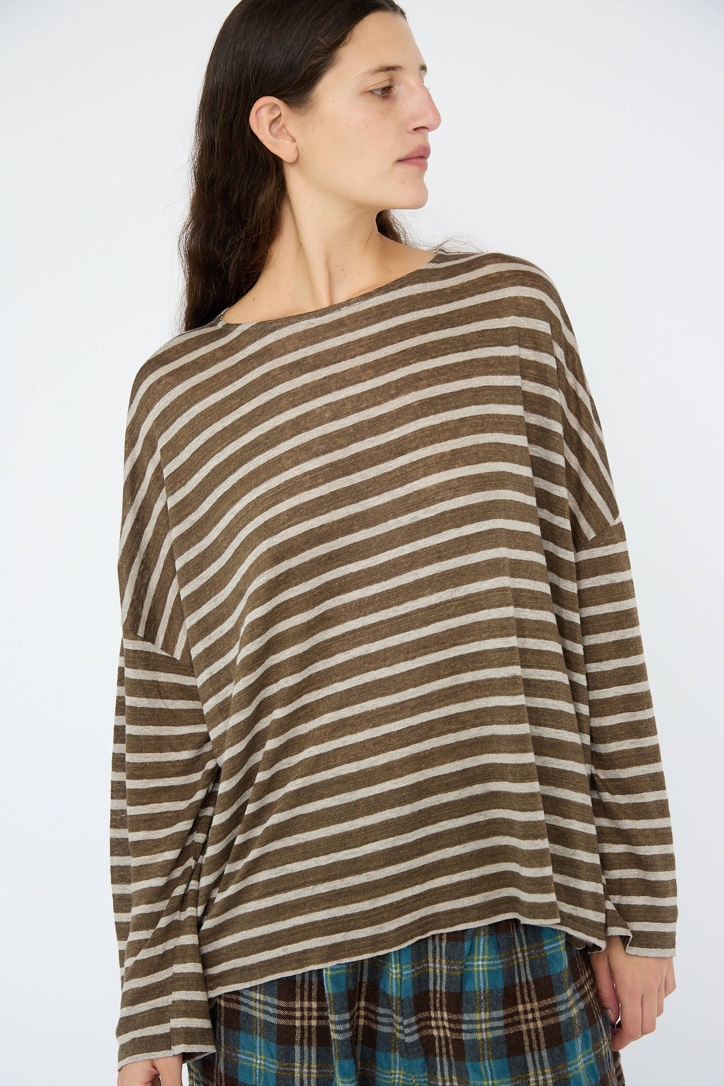 Wearing the Linen Stripe Pullover in Khaki and Natural by Antiquités Noir, the individual pairs it with plaid pants and gazes off to the side against a plain background.