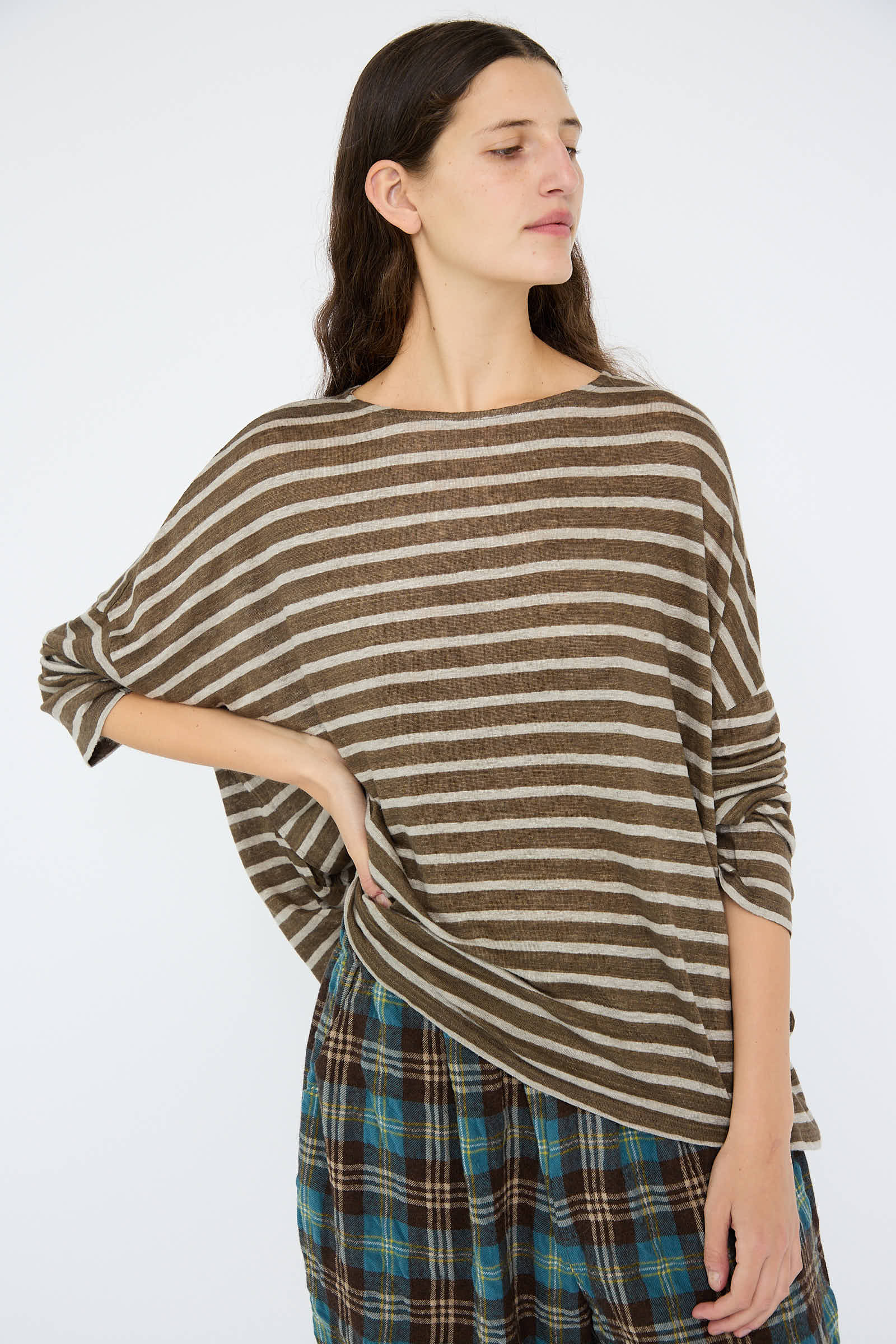 A person in a Linen Stripe Pullover in Khaki and Natural by Antiquités Noir, paired with plaid pants, stands with hand on hip against a plain background.