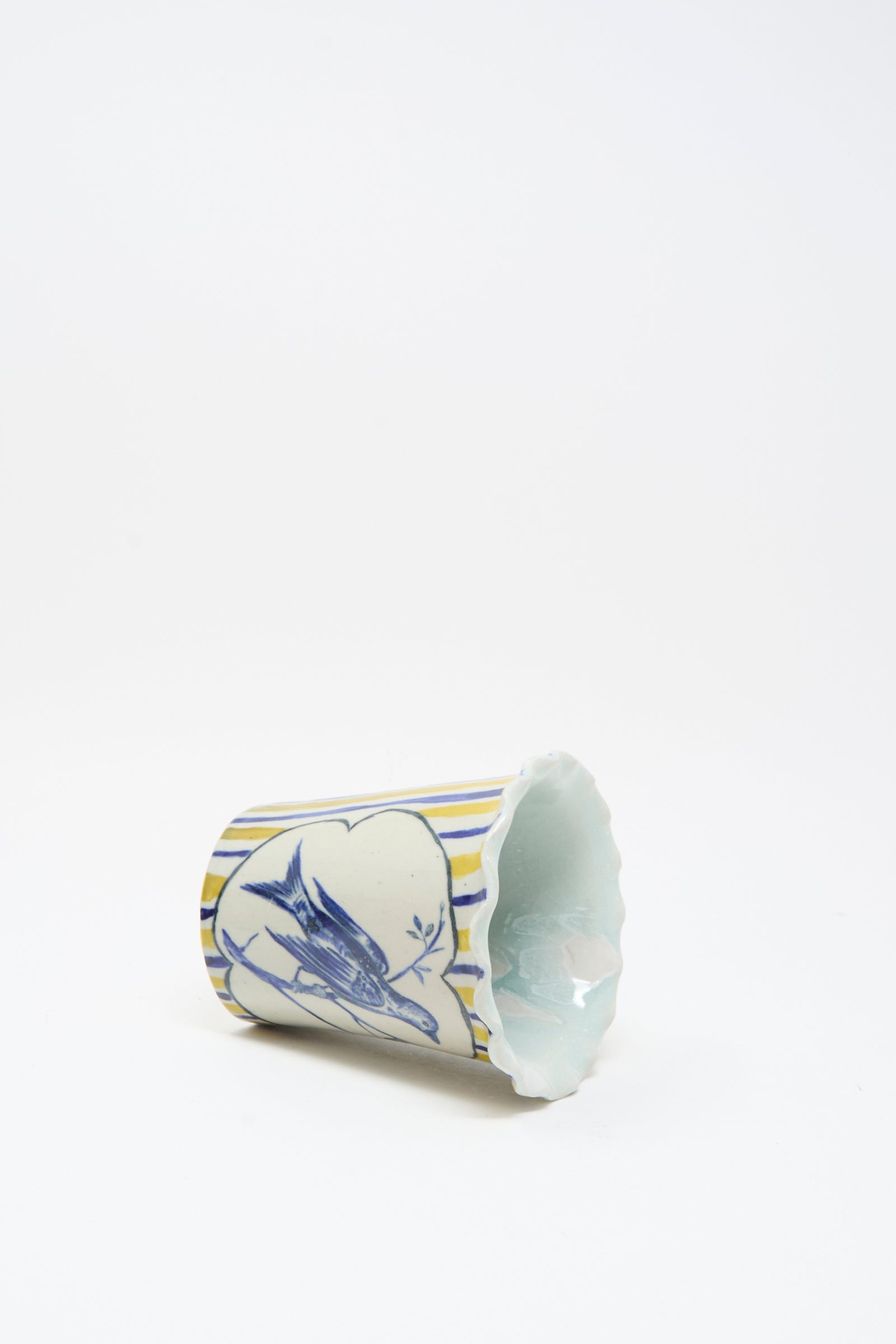 The Hand Painted Bird Vessel by Ariel Clute is a ceramic cup with scalloped edges, blue bird illustrations, and yellow stripes on a white background, handmade in California.