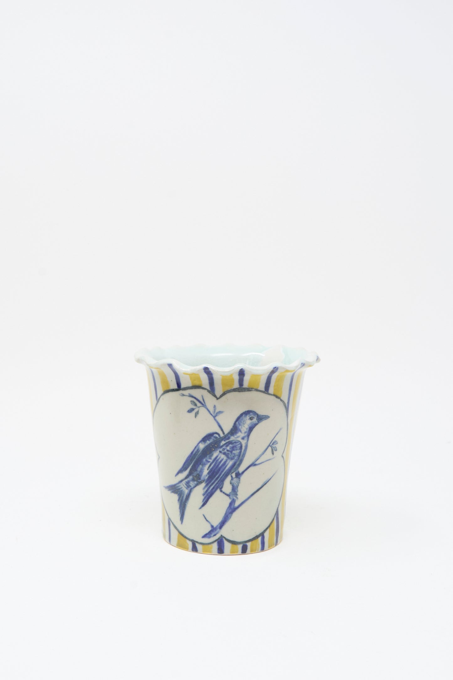 The Ariel Clute Hand Painted Bird Vessel is a white ceramic cup with blue and yellow stripes, featuring a hand painted porcelain bird inside a heart-shaped frame on the front.