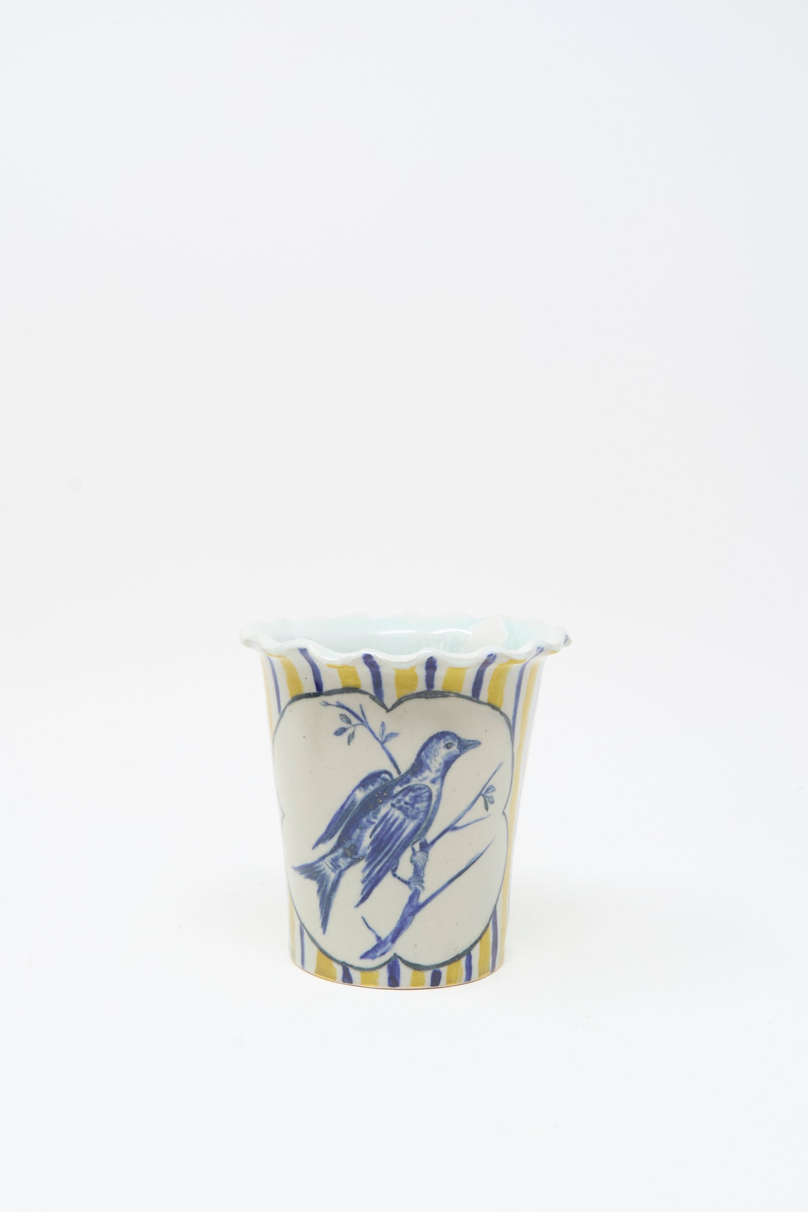 The Ariel Clute Hand Painted Bird Vessel is a white ceramic cup with blue and yellow stripes, featuring a hand painted porcelain bird inside a heart-shaped frame on the front.