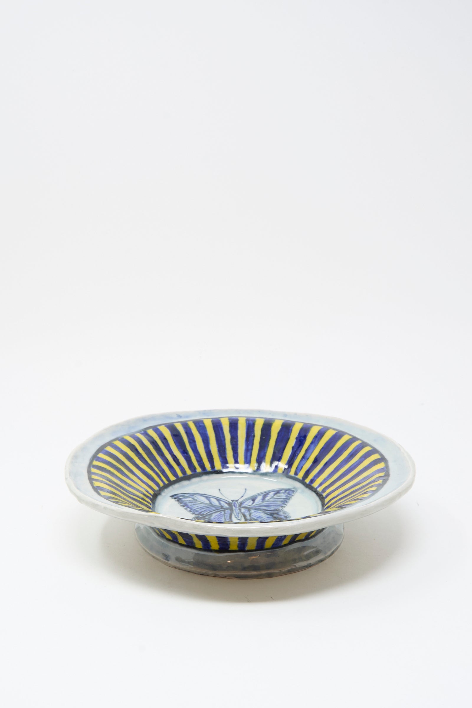 The Ariel Clute Hand Painted Butterfly Bowl features blue and yellow radial stripes with a blue butterfly design at the center, handmade in California and displayed on a plain white background.