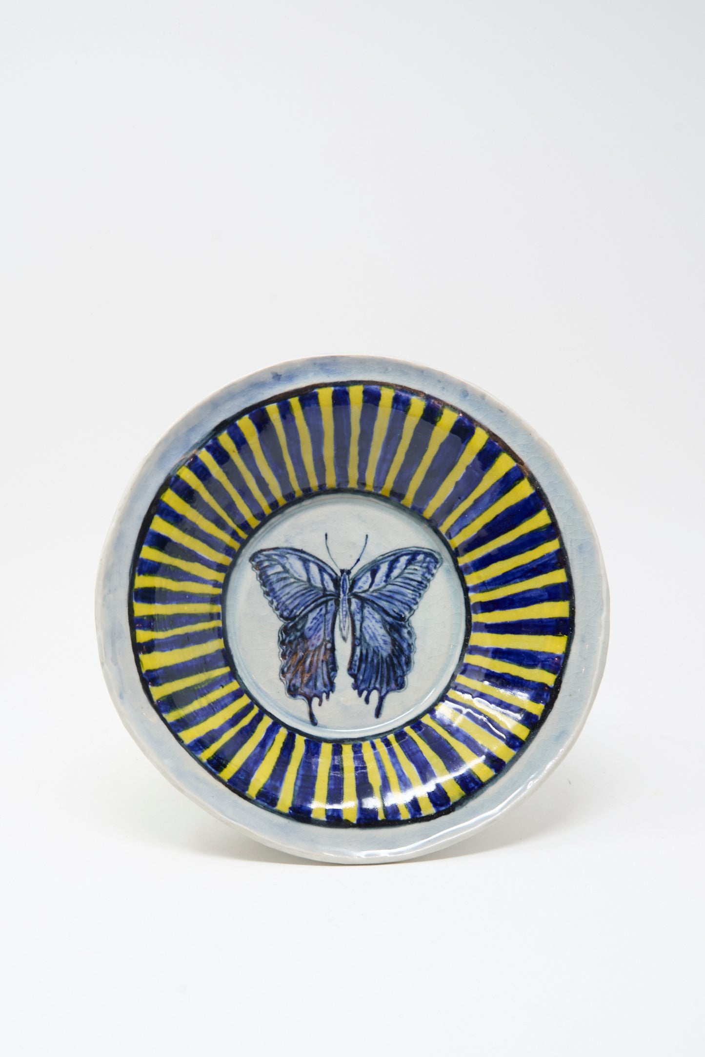 The Ariel Clute Hand Painted Butterfly Bowl is a handmade ceramic piece from California, featuring a blue butterfly at the center with alternating blue and yellow radial stripes on a white background.