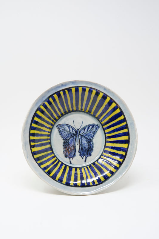 The Ariel Clute Hand Painted Butterfly Bowl is a handmade ceramic piece from California, featuring a blue butterfly at the center with alternating blue and yellow radial stripes on a white background.