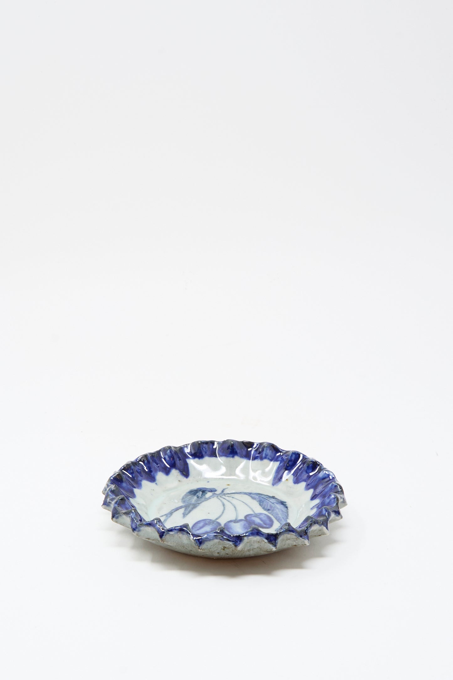 The Rustic Cherries Plate by Ariel Clute is a handmade, shallow round ceramic dish from California with a ruffled blue rim and delicate blue floral patterns on white.