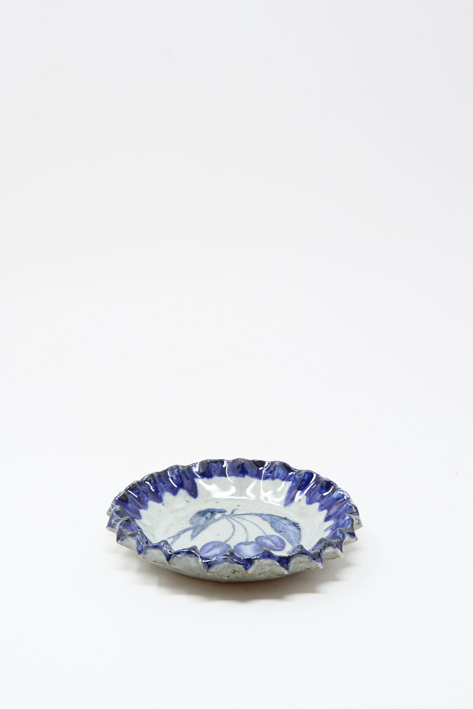 The Rustic Cherries Plate by Ariel Clute is a handmade, shallow round ceramic dish from California with a ruffled blue rim and delicate blue floral patterns on white.