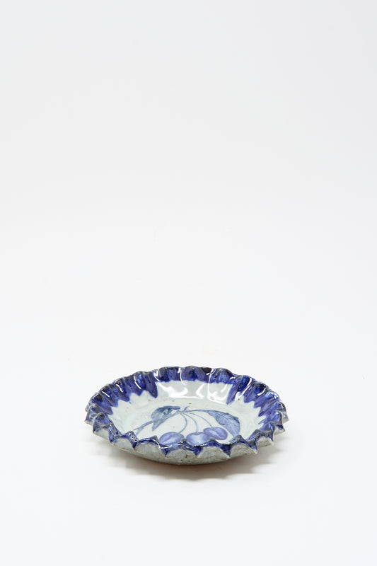 The Rustic Cherries Plate by Ariel Clute is a handmade, shallow round ceramic dish from California with a ruffled blue rim and delicate blue floral patterns on white.