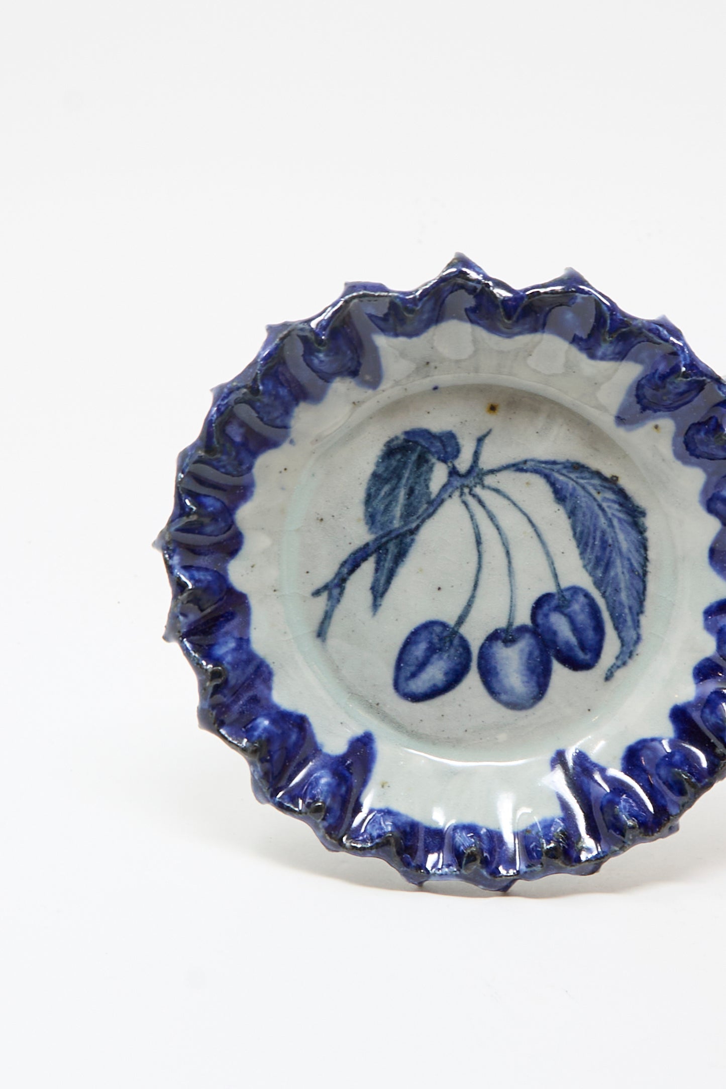 The Rustic Cherries Plate by Ariel Clute is a hand-painted porcelain piece, featuring a ruffled blue edge and a cherry design, handmade in California.