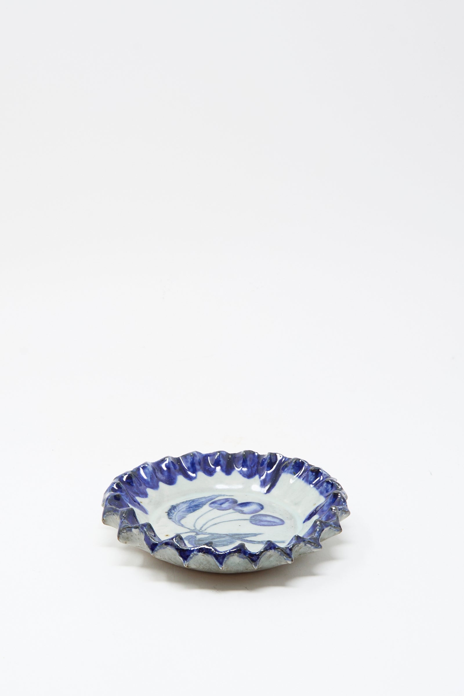 The Rustic Cherries Plate by Ariel Clute is a small, round ceramic dish with a ruffled edge and blue glaze accents on a white background, handmade in California.