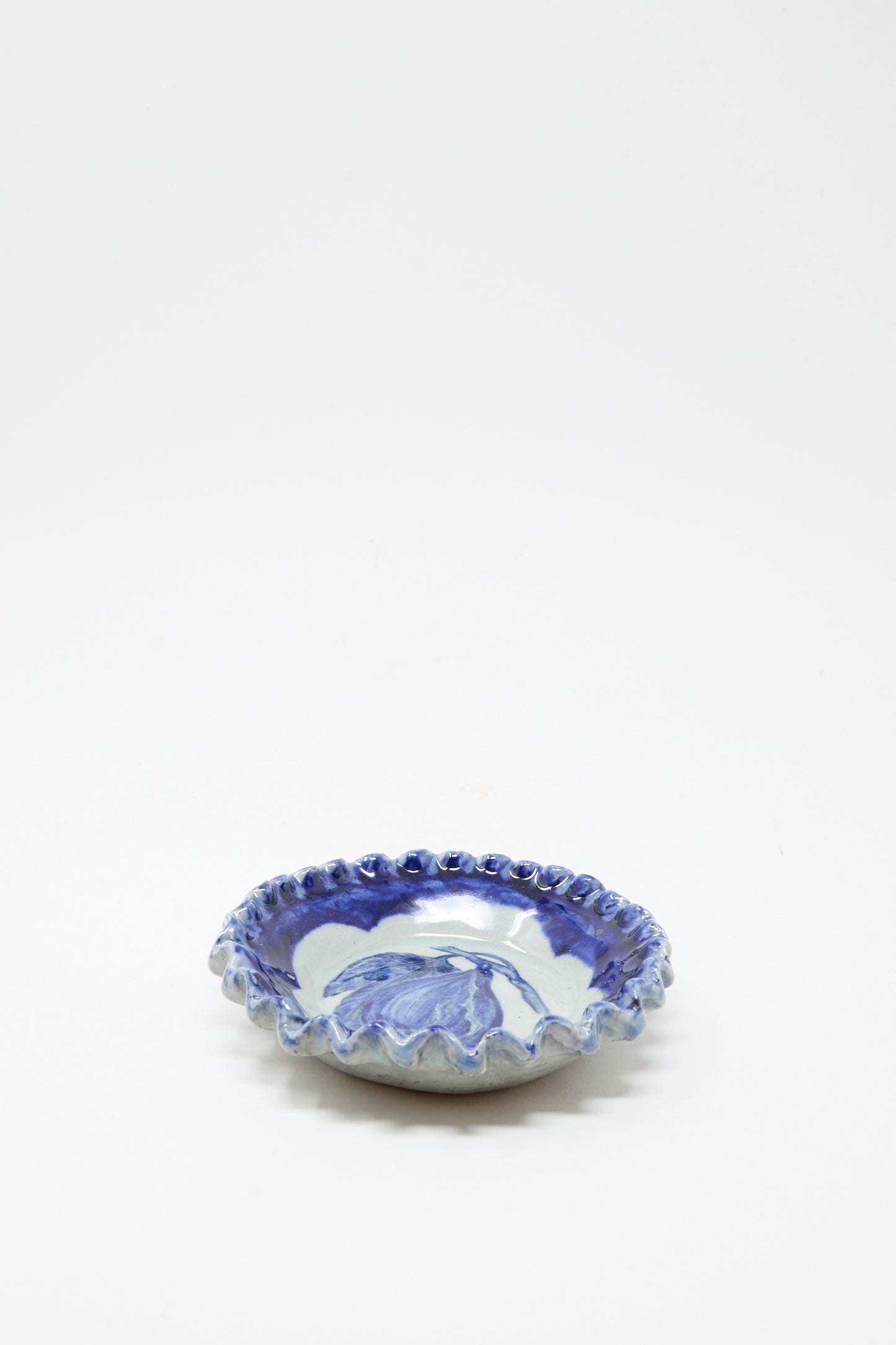 The Ariel Clute Rustic Fig Plate is a shallow, round ceramic dish with a scalloped edge, hand painted with blue floral patterns on a white porcelain background.